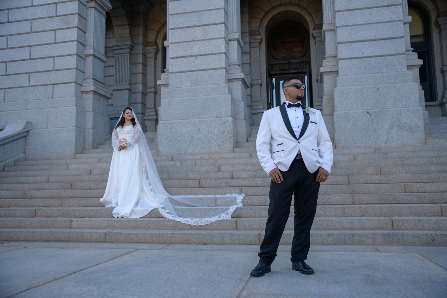 Bride in white gown with veil and groom in white tuxedo posing on stone steps outside a grand building.