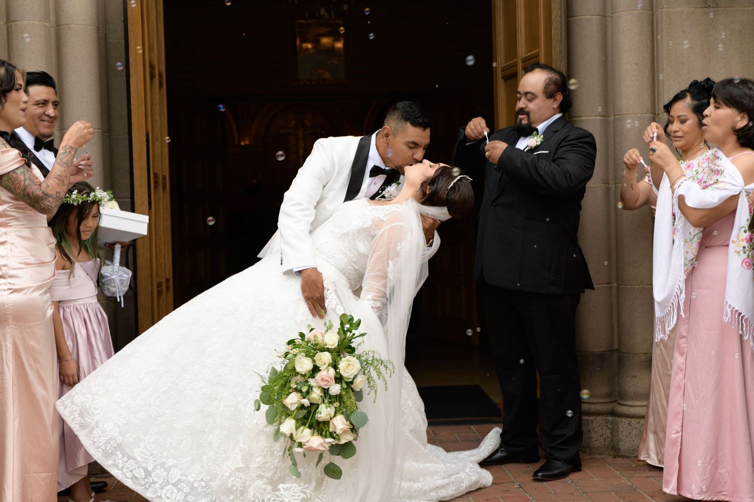 Newlywed couple sharing a romantic dip kiss outside church doors, surrounded by wedding party in blush pink gowns.