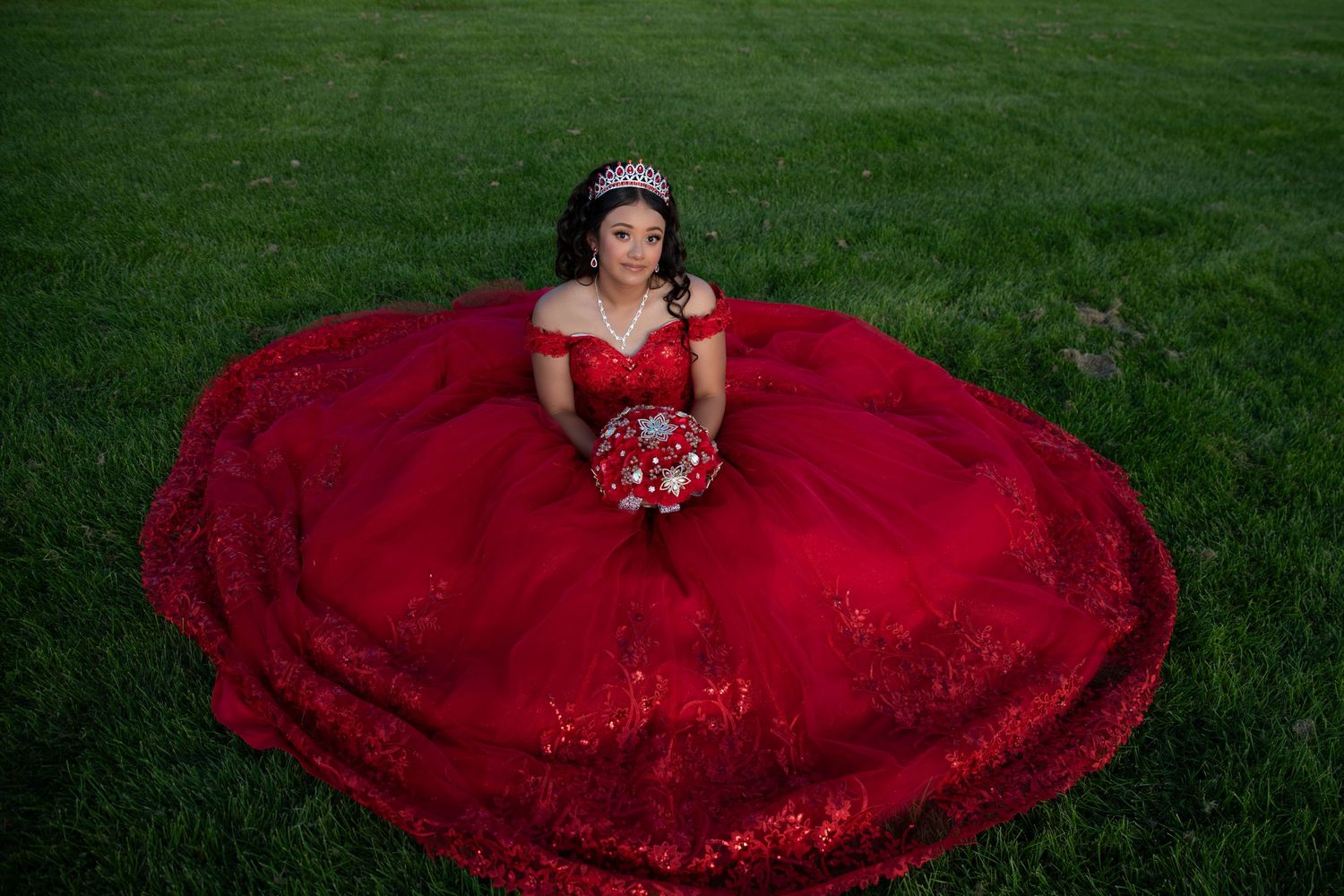 Aerial view of young woman in red sequined quinceañera dress sitting on lush green grass lawn.