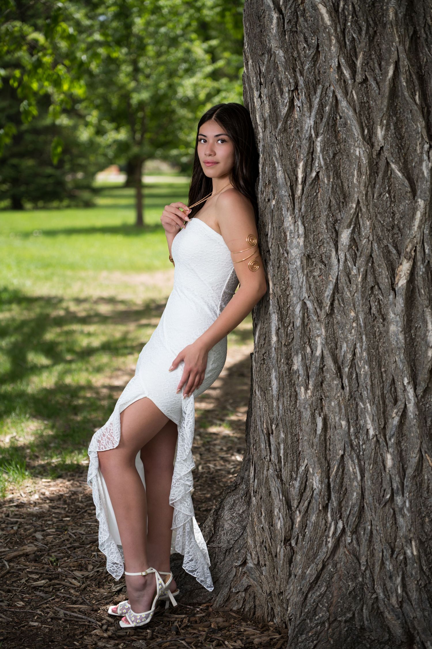 Young woman in white lace high-low dress leans gracefully against large tree trunk in sunny park setting.