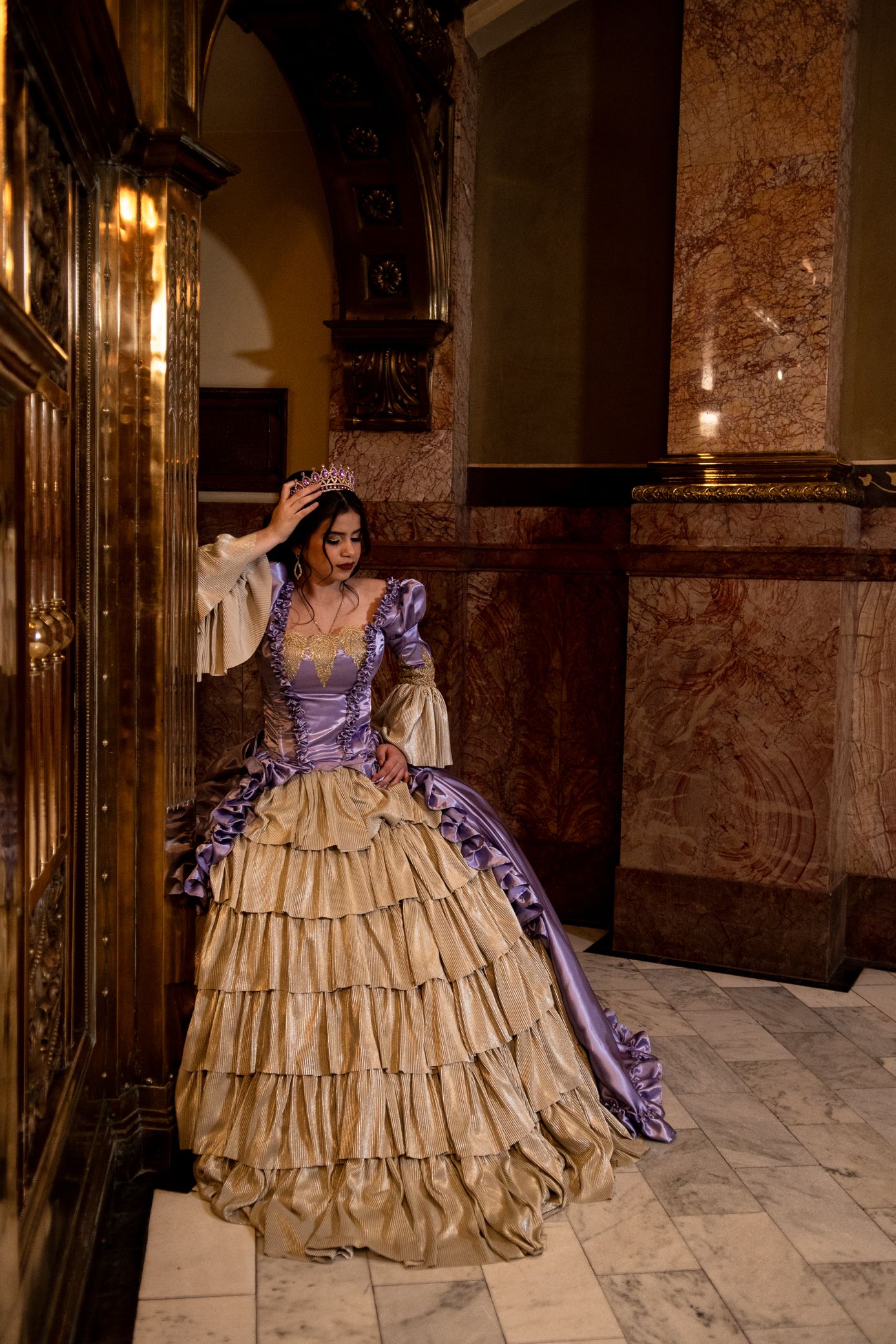 Woman in lavender Victorian ball gown with tiara posing beside wooden door in ornate marble hallway.