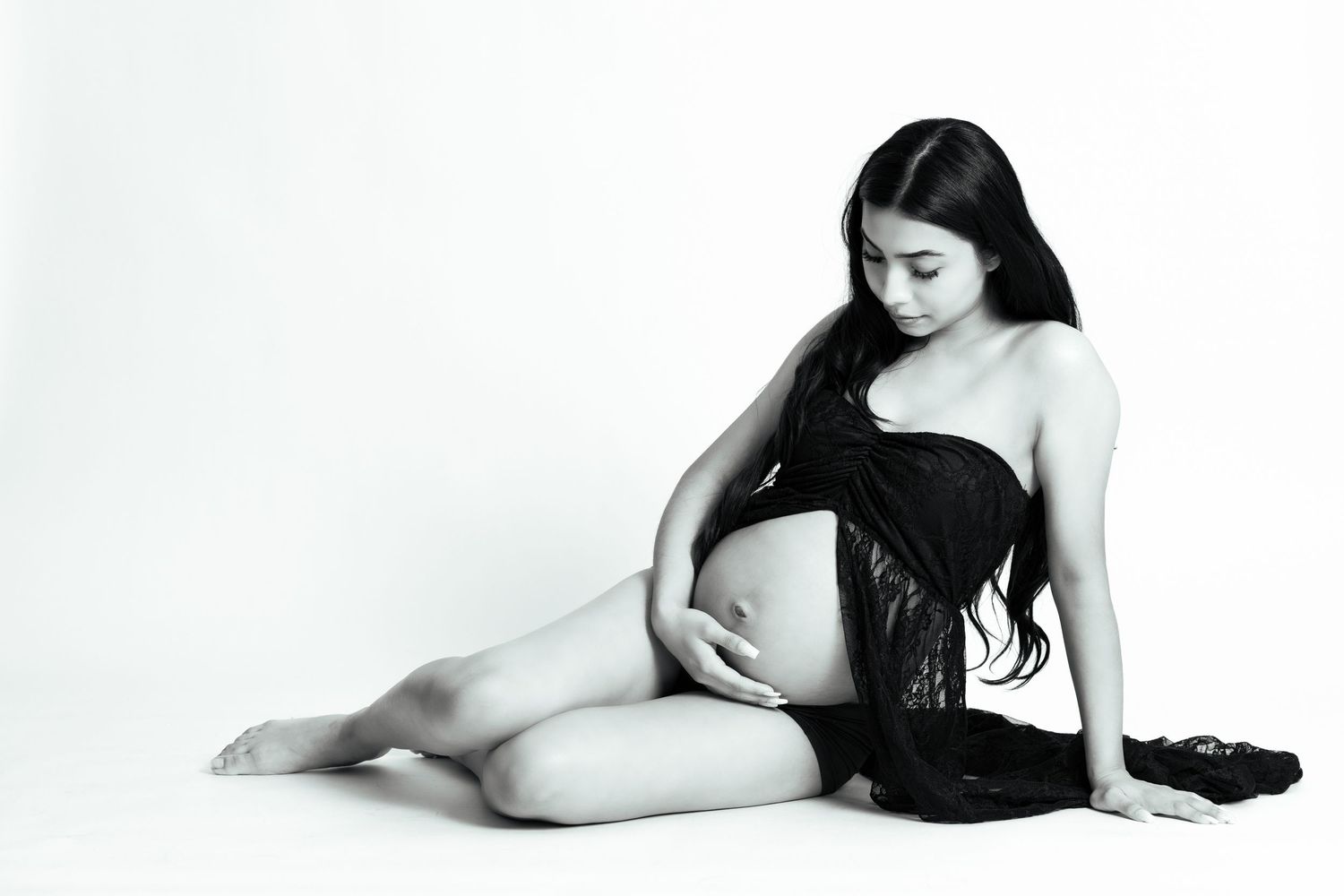 Black and white maternity photo of a pregnant woman sitting gracefully, cradling her baby bump in a studio setting.