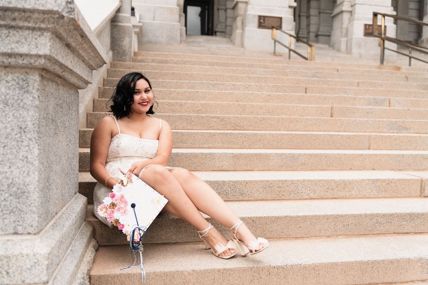 Young woman in white dress sitting on stone steps holding a floral graduation cap, smiling outdoors.