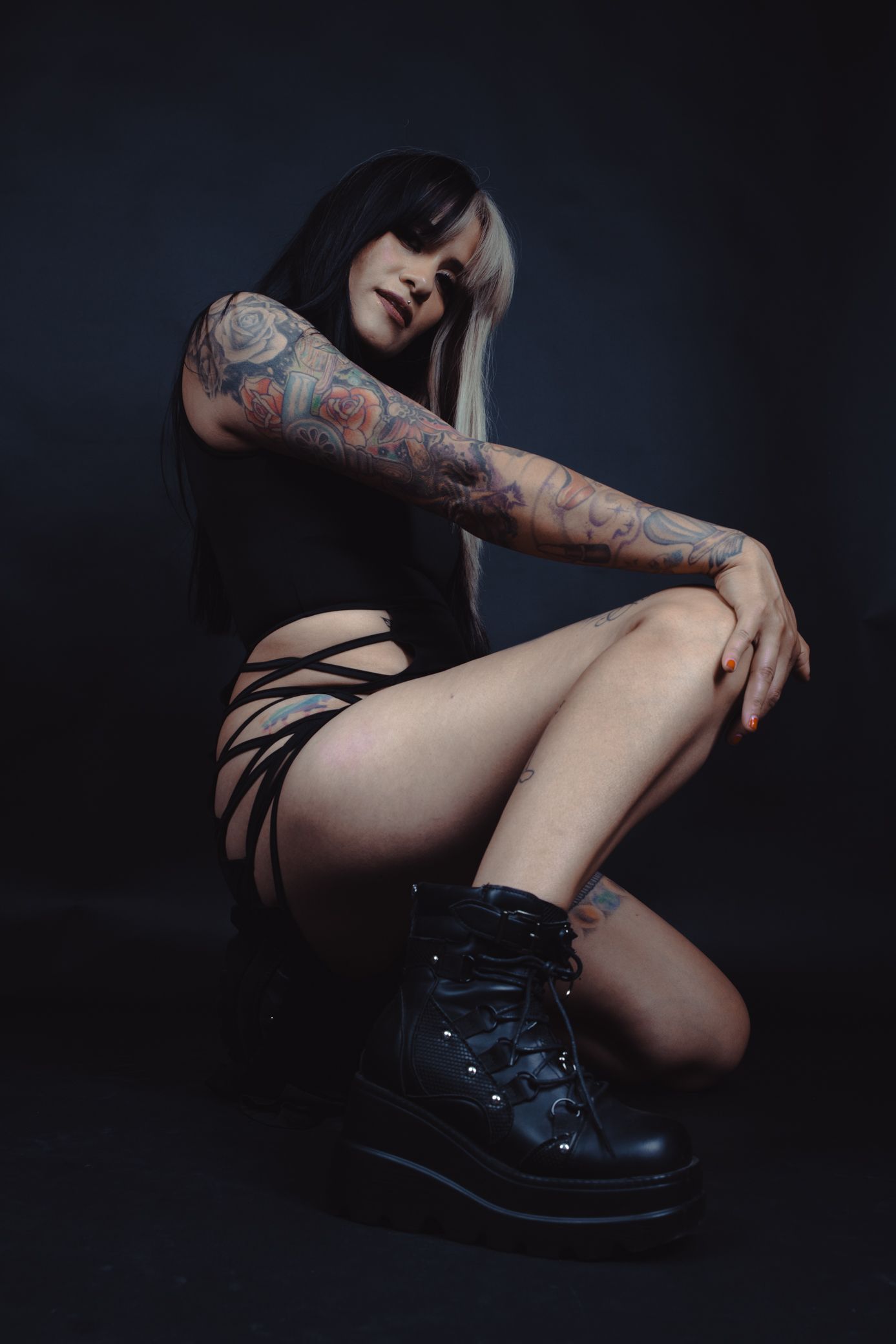 Tattooed woman in black outfit with chunky boots posing on dark studio background.