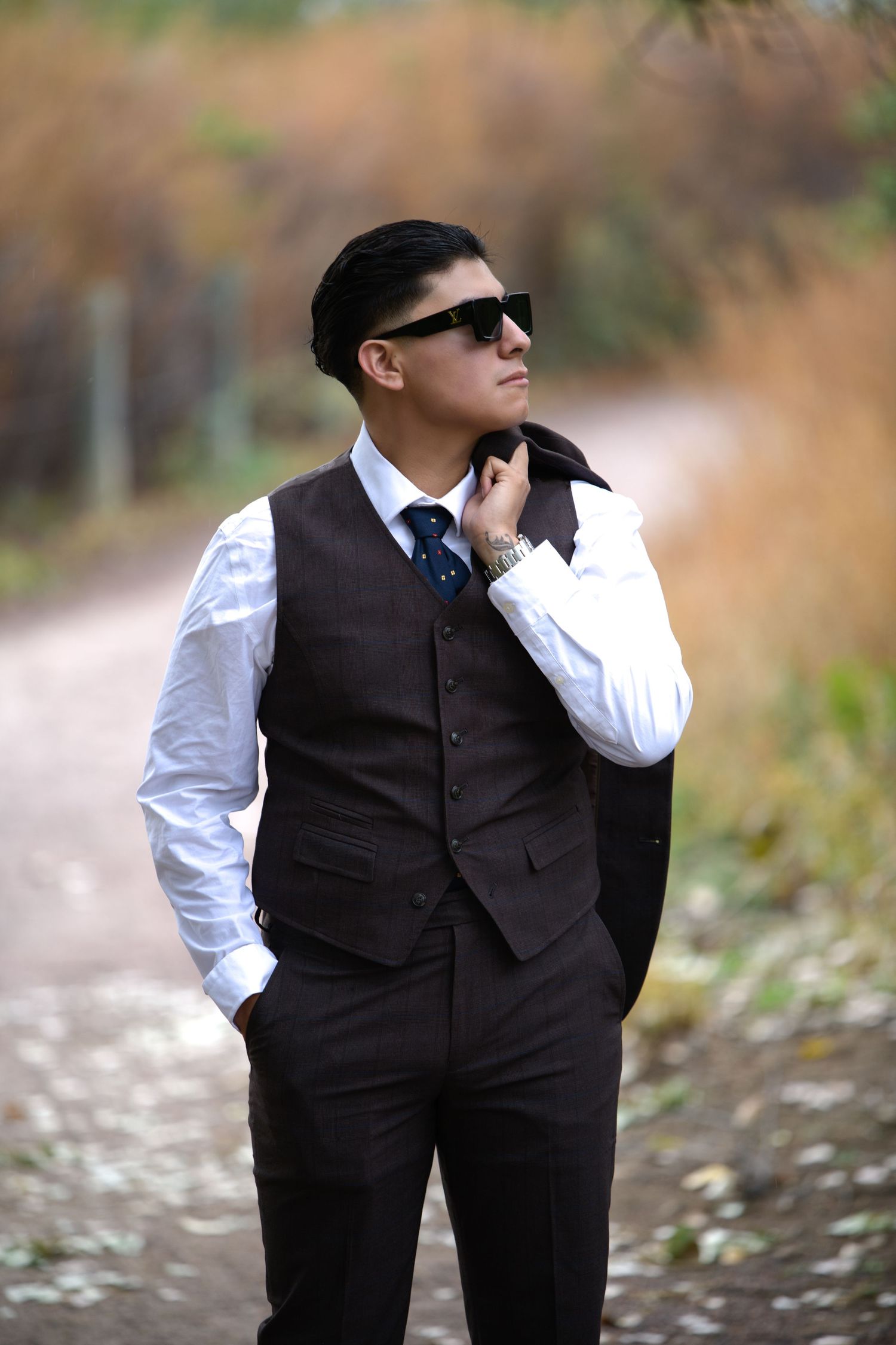 Stylish man in dark vest, white shirt, blue tie, and sunglasses posing on a nature path.