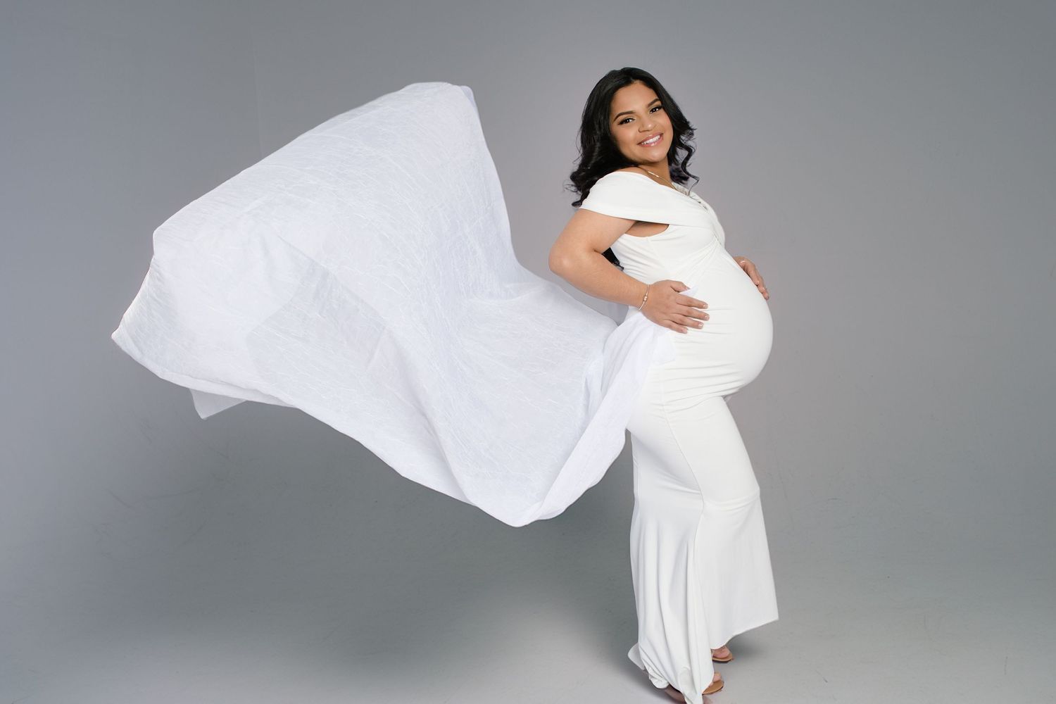 A smiling pregnant woman in a white fitted gown poses with flowing fabric against a gray studio backdrop.