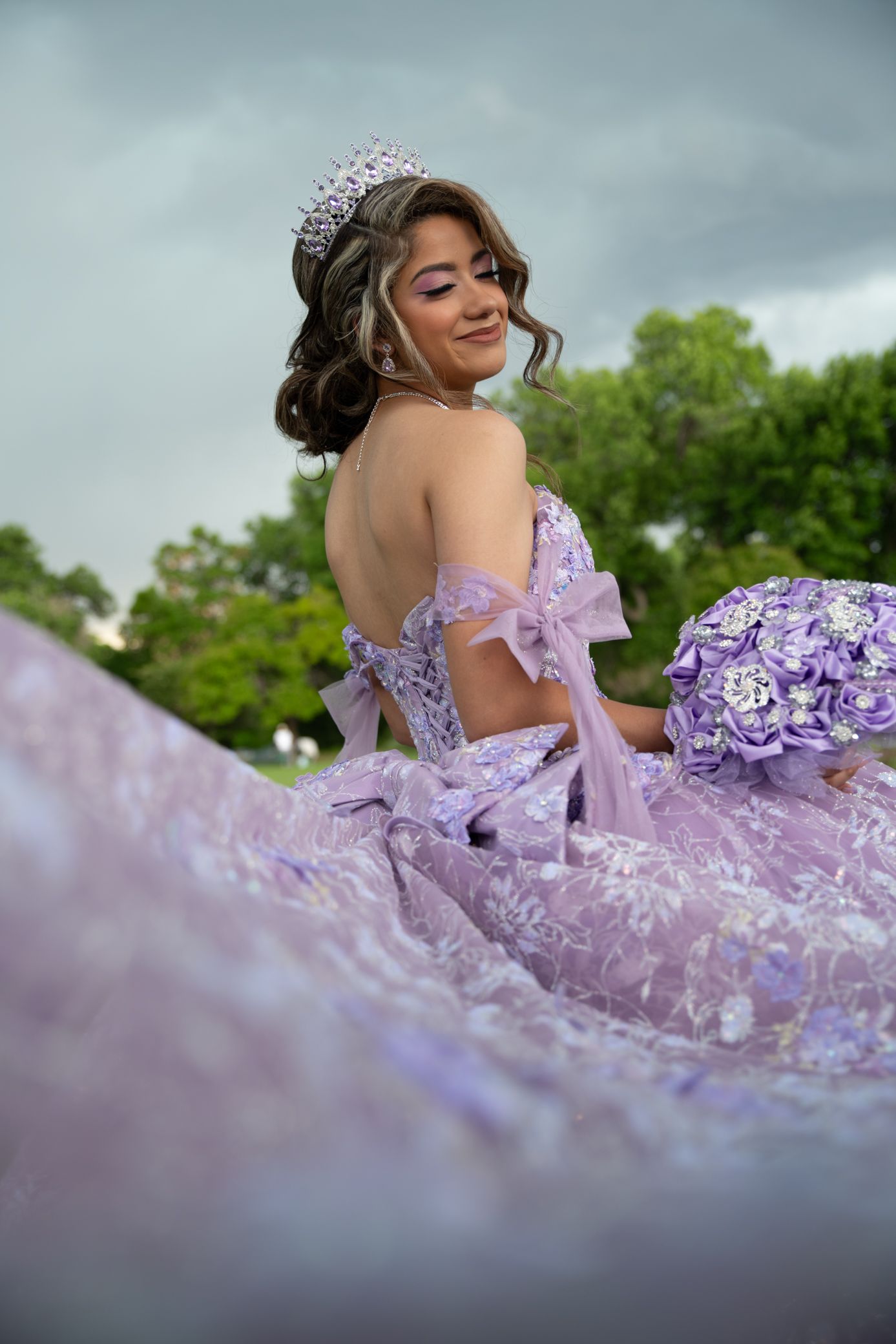 Girl in purple quinceañera dress with tiara sitting outdoors holding floral bouquet.