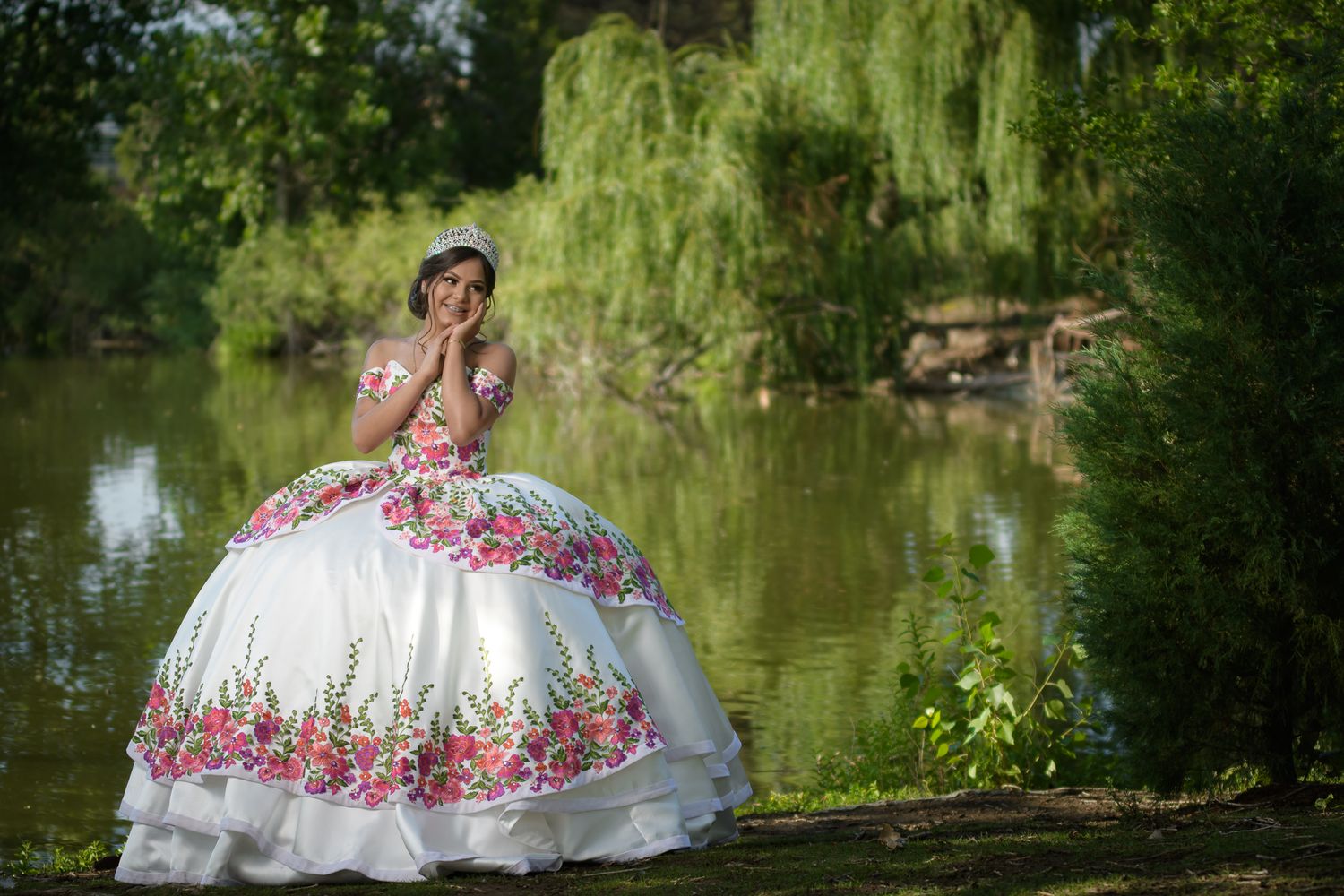 Quinceañera in white floral embroidered ball gown posing near a serene pond outdoors.