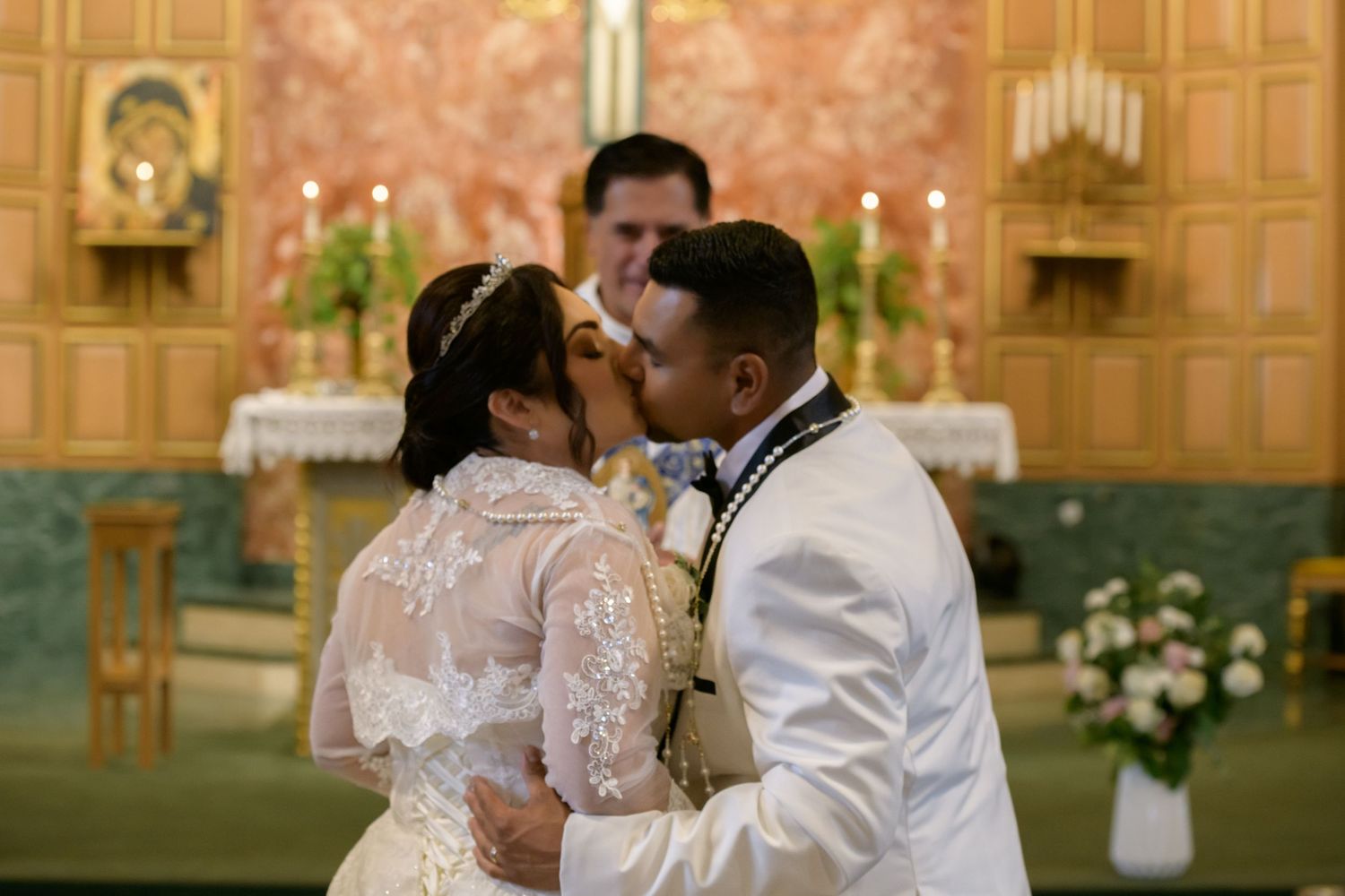 Bride and groom share first kiss at church altar during wedding ceremony, bride in lace gown with tiara.