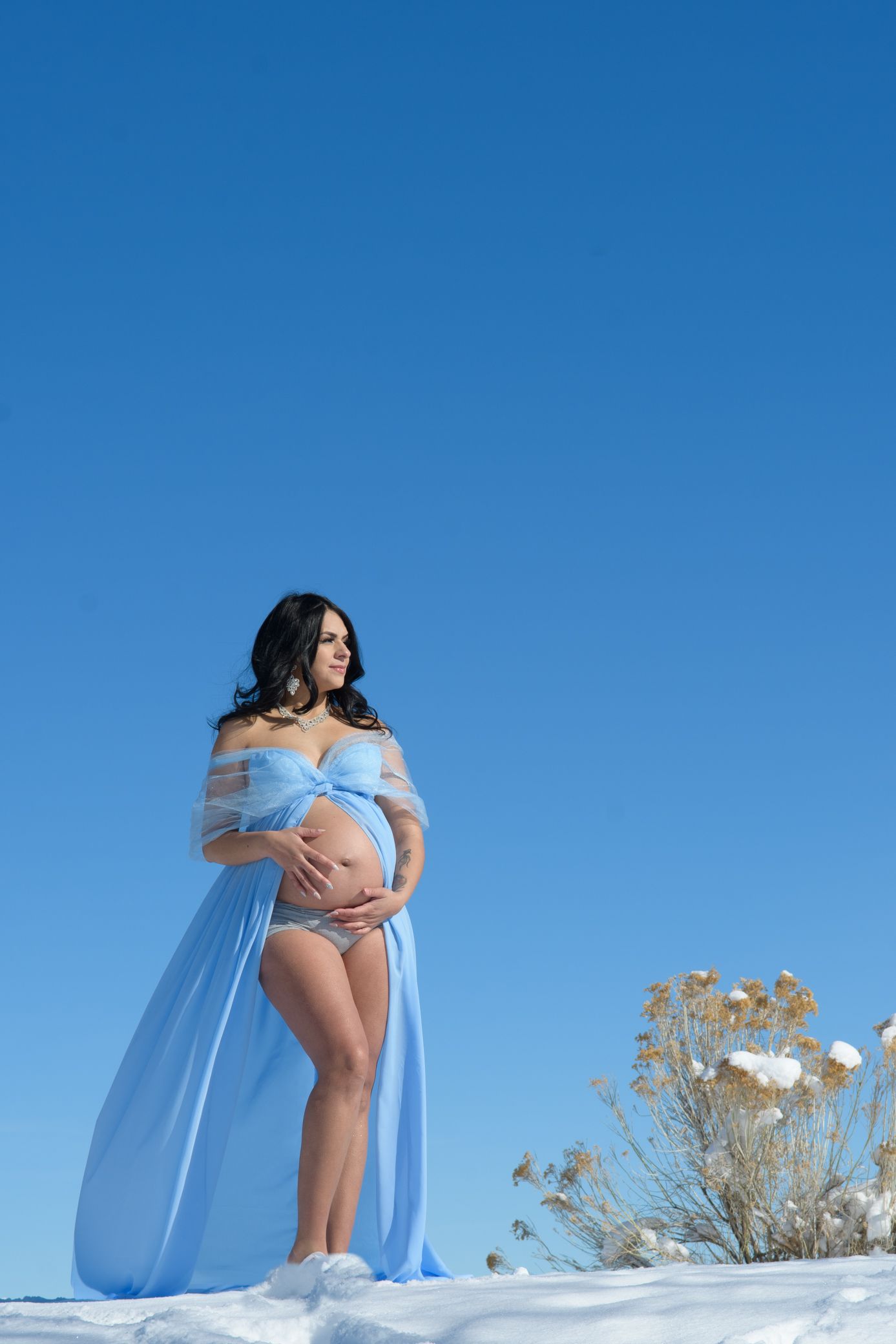 Pregnant woman in flowing blue sheer cape standing in snowy winter landscape under clear blue sky.