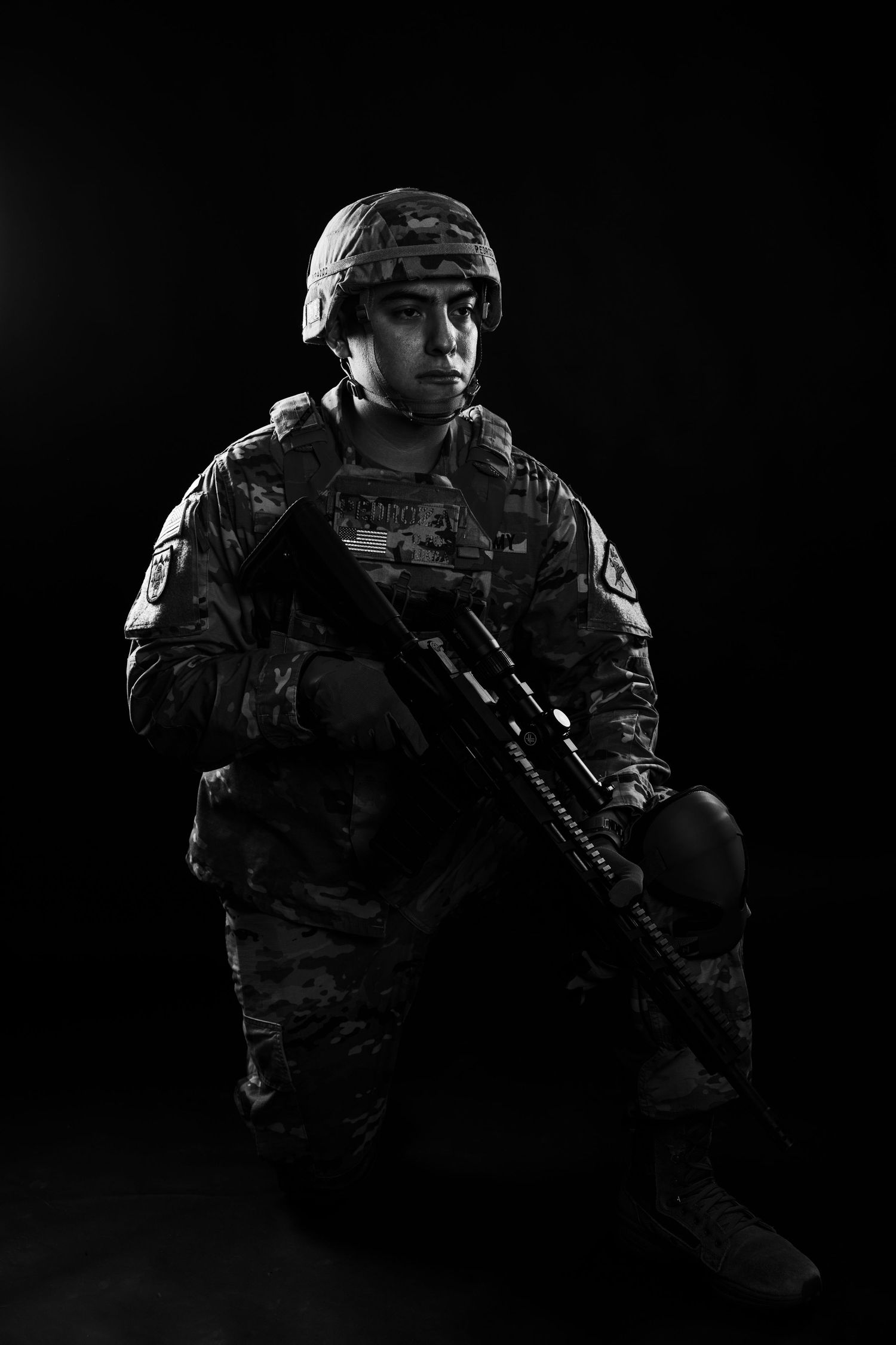 Black and white portrait of female US Army soldier in full combat uniform kneeling with rifle on dark background.
