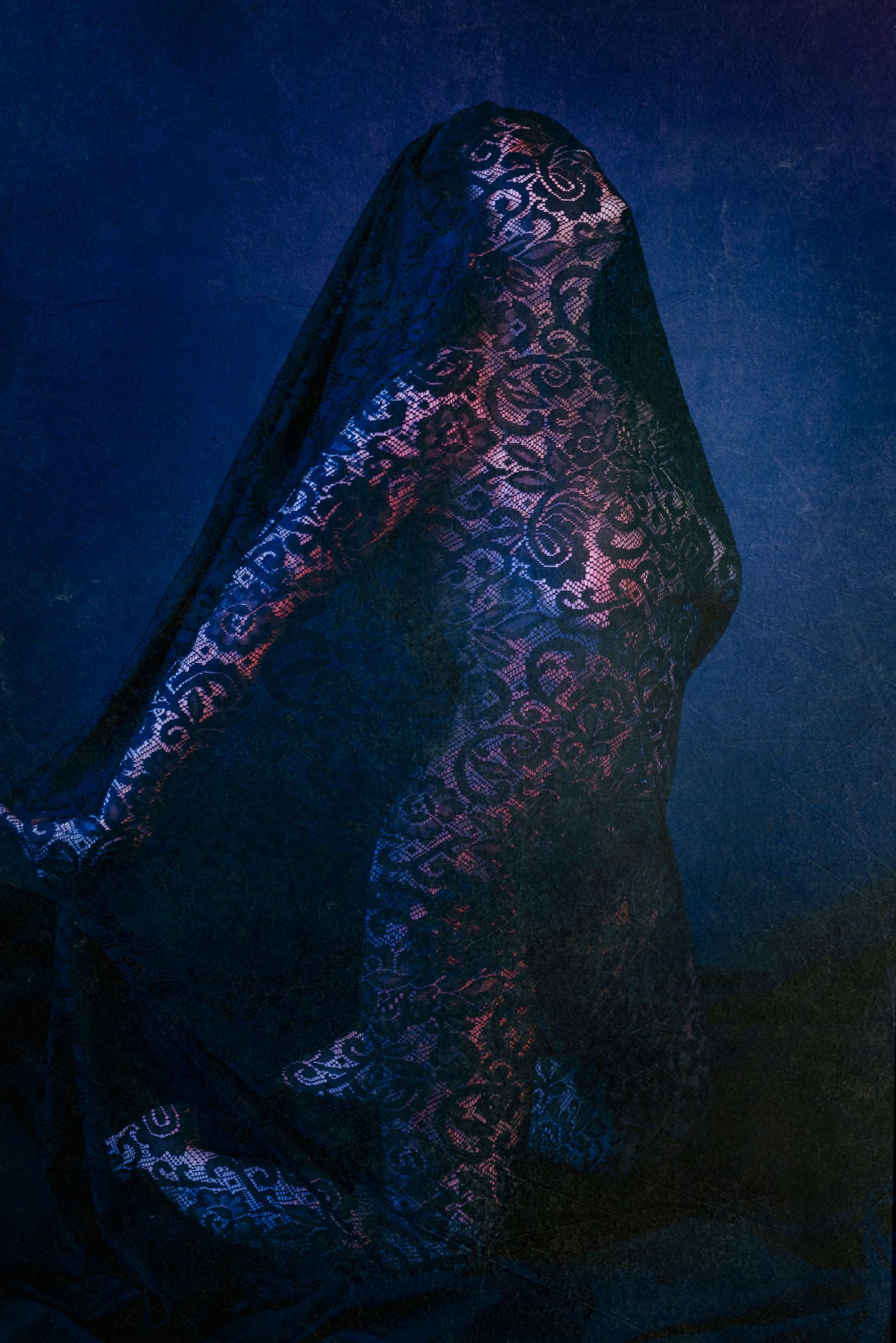 A figure draped in dark floral lace fabric against a deep blue textured background in moody artistic photography.