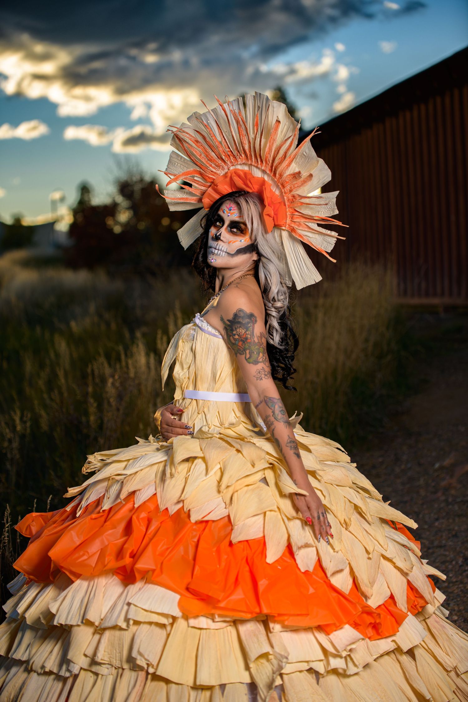 Woman in elaborate orange and cream ruffled gown with sugar skull makeup and sunburst headdress at sunset.