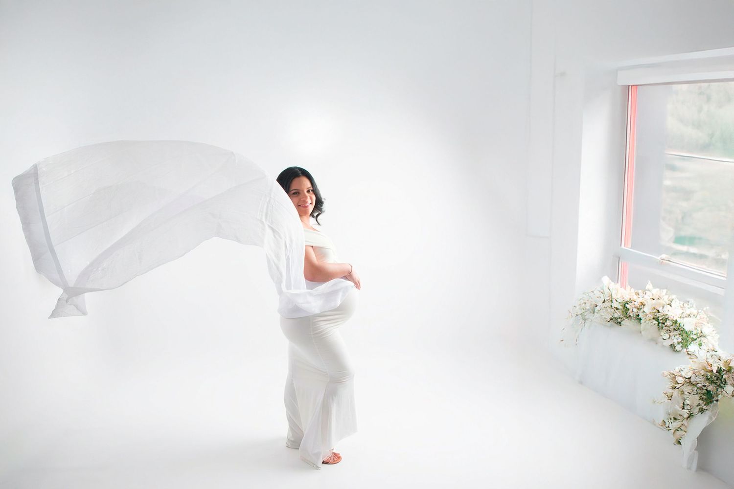 Pregnant woman in white dress with flowing fabric in a bright white studio maternity photo.