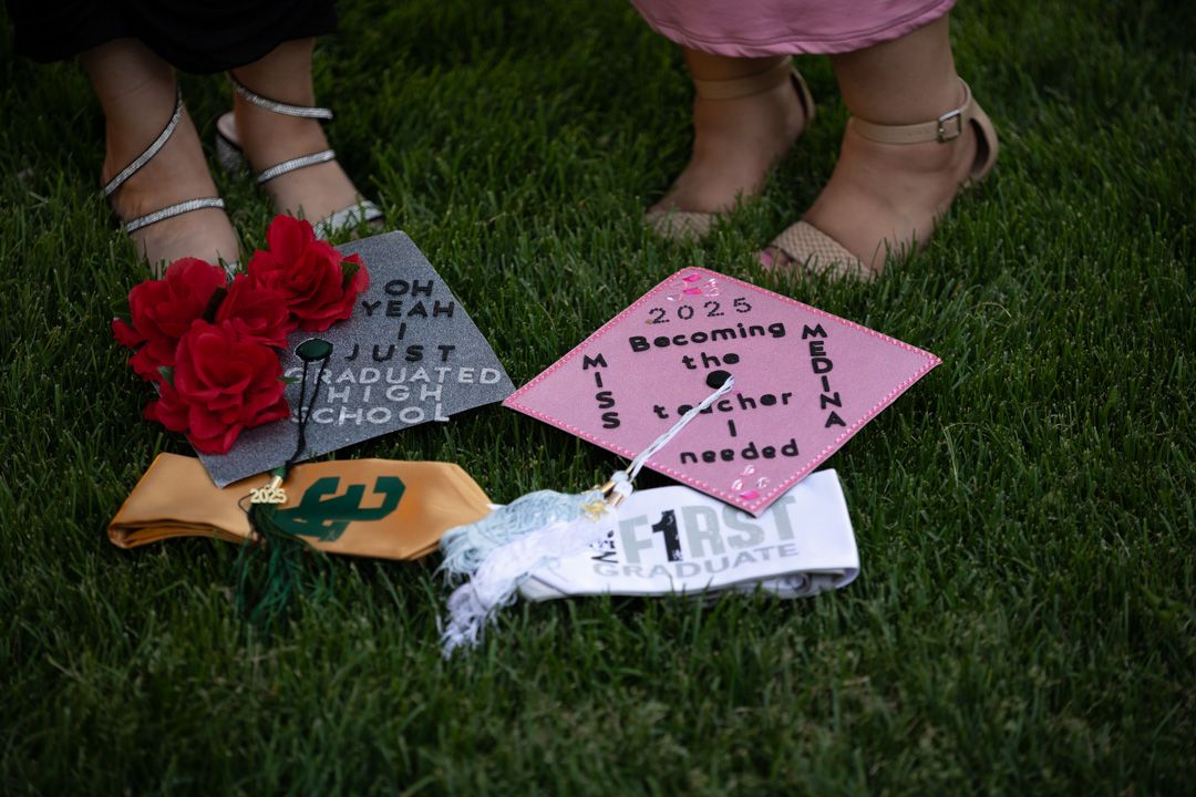 Two graduates standing on grass with decorated caps reading teacher-themed messages and red flowers.
