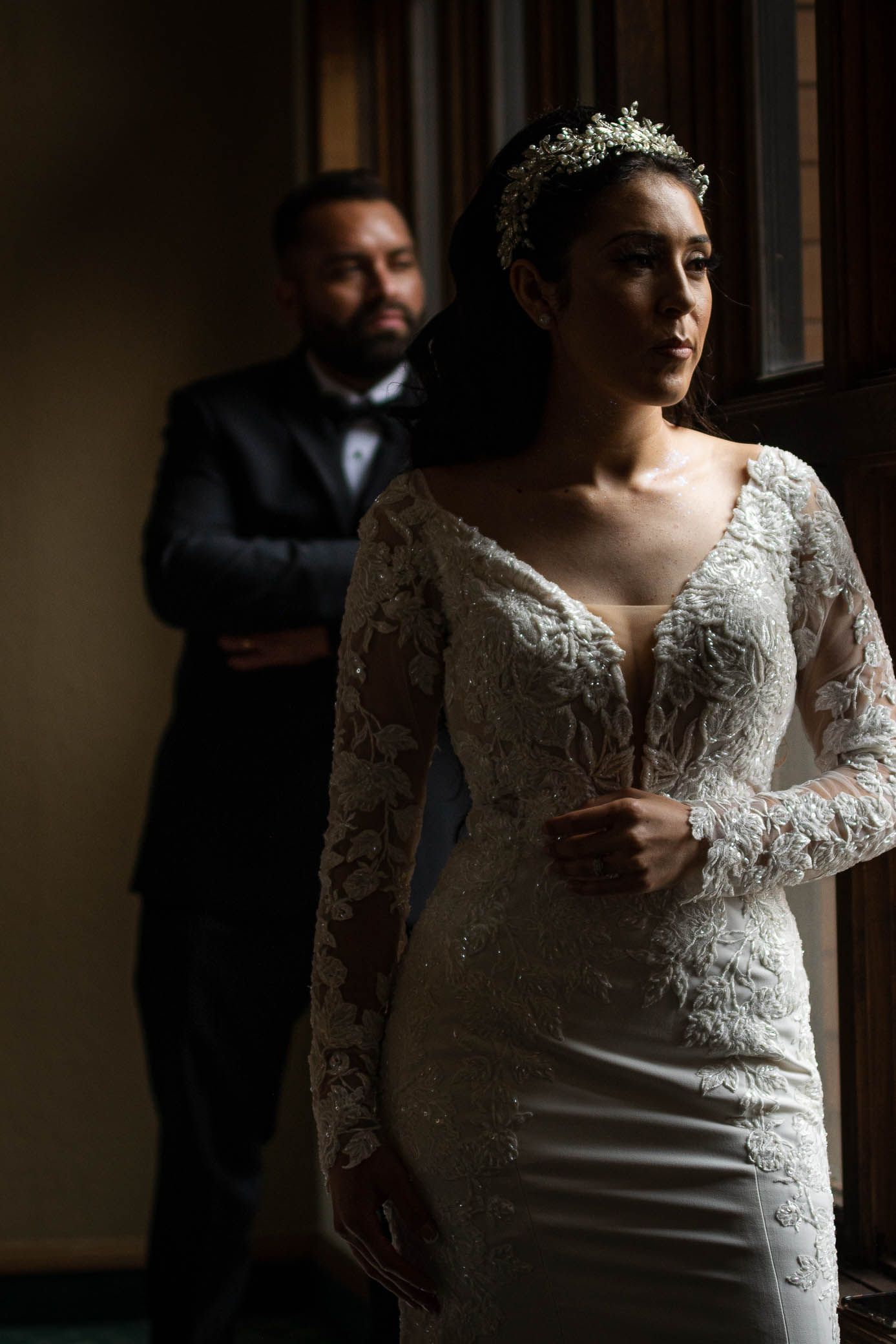 Bride in long-sleeve lace wedding gown stands by window as groom watches from the shadows.