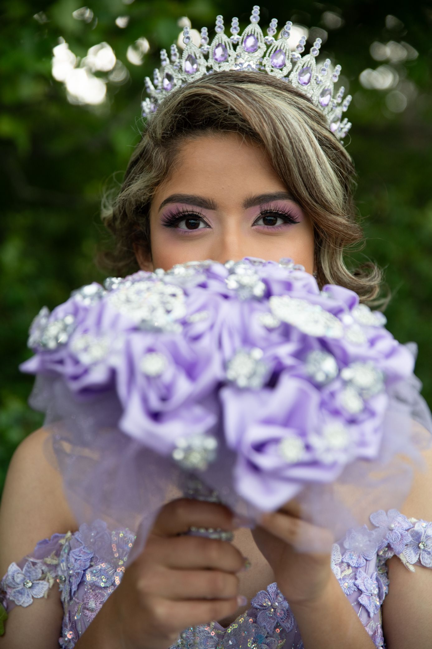 Quinceañera girl hiding behind purple rose bouquet wearing crystal tiara and lavender gown.