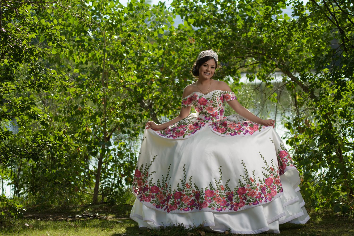 Smiling young woman twirls white floral embroidered quinceañera ball gown among lush green trees.