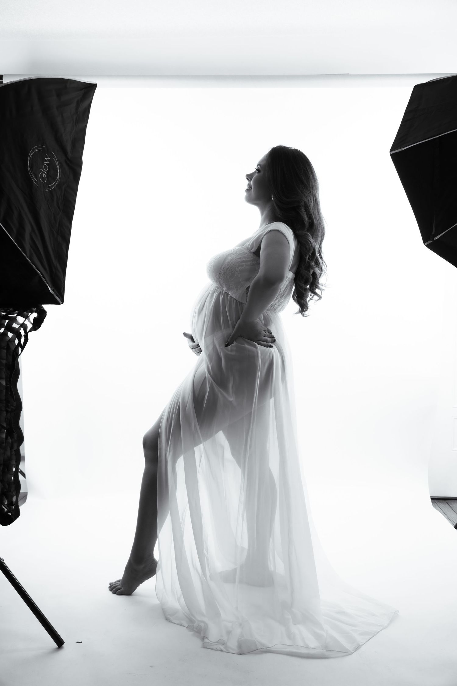 Silhouette of pregnant woman in sheer white gown posed between studio softbox lights.