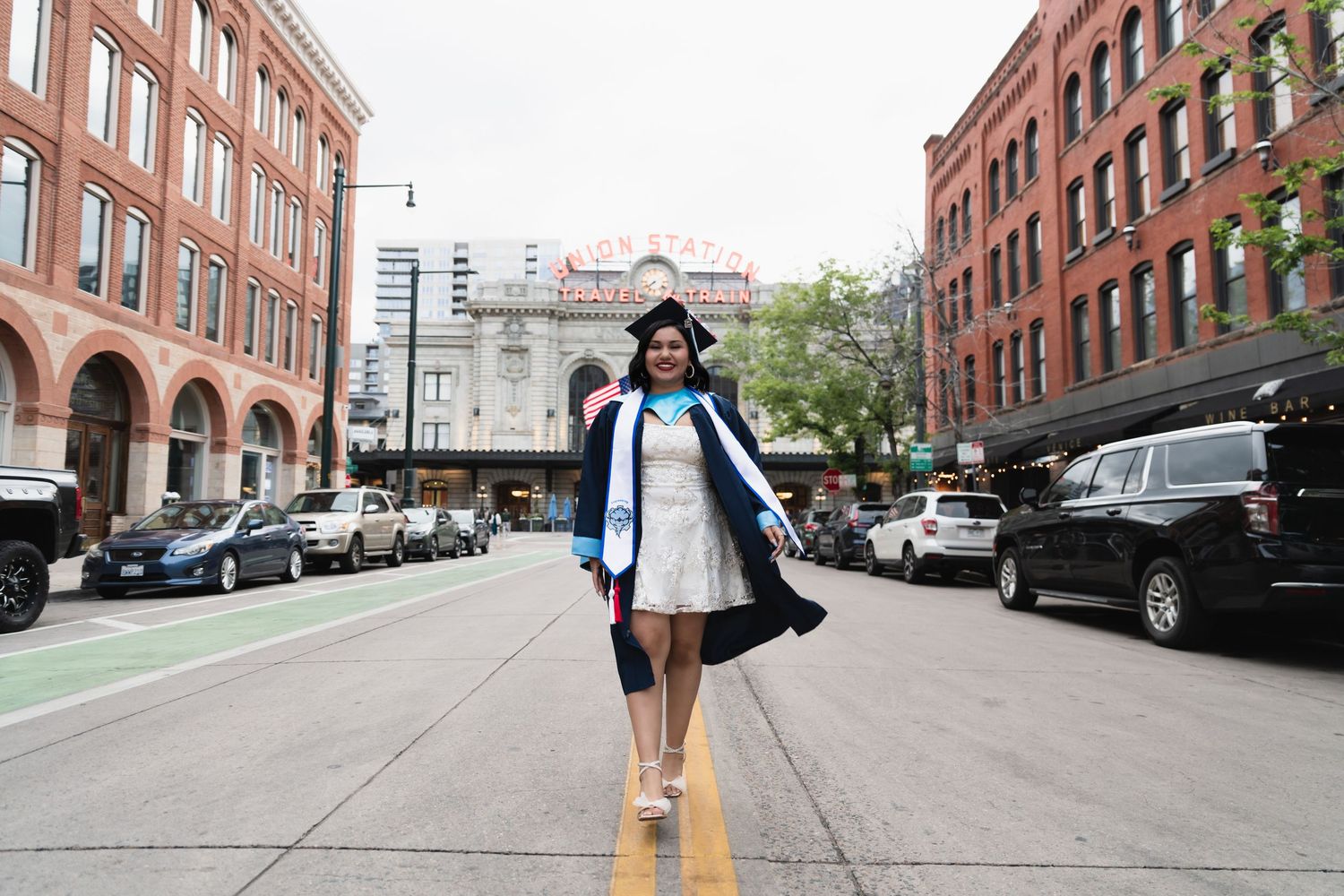 Smiling graduate in cap and gown walking on urban street in white dress during graduation celebration.