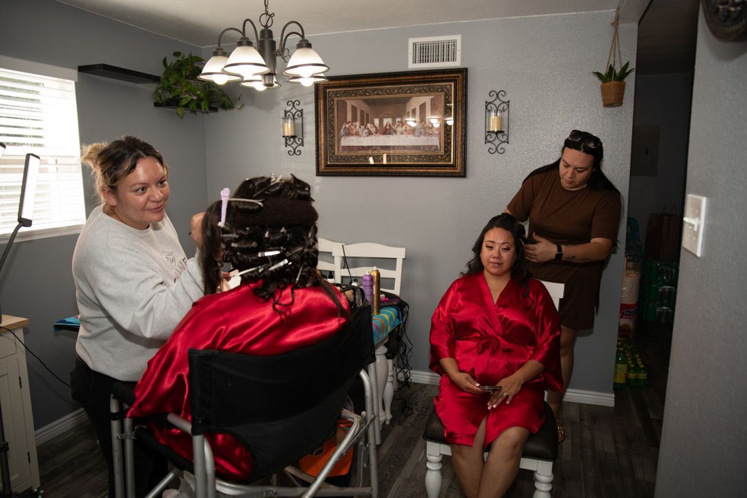 Two women getting hair and makeup done in a home salon setting with a chandelier and Last Supper painting.