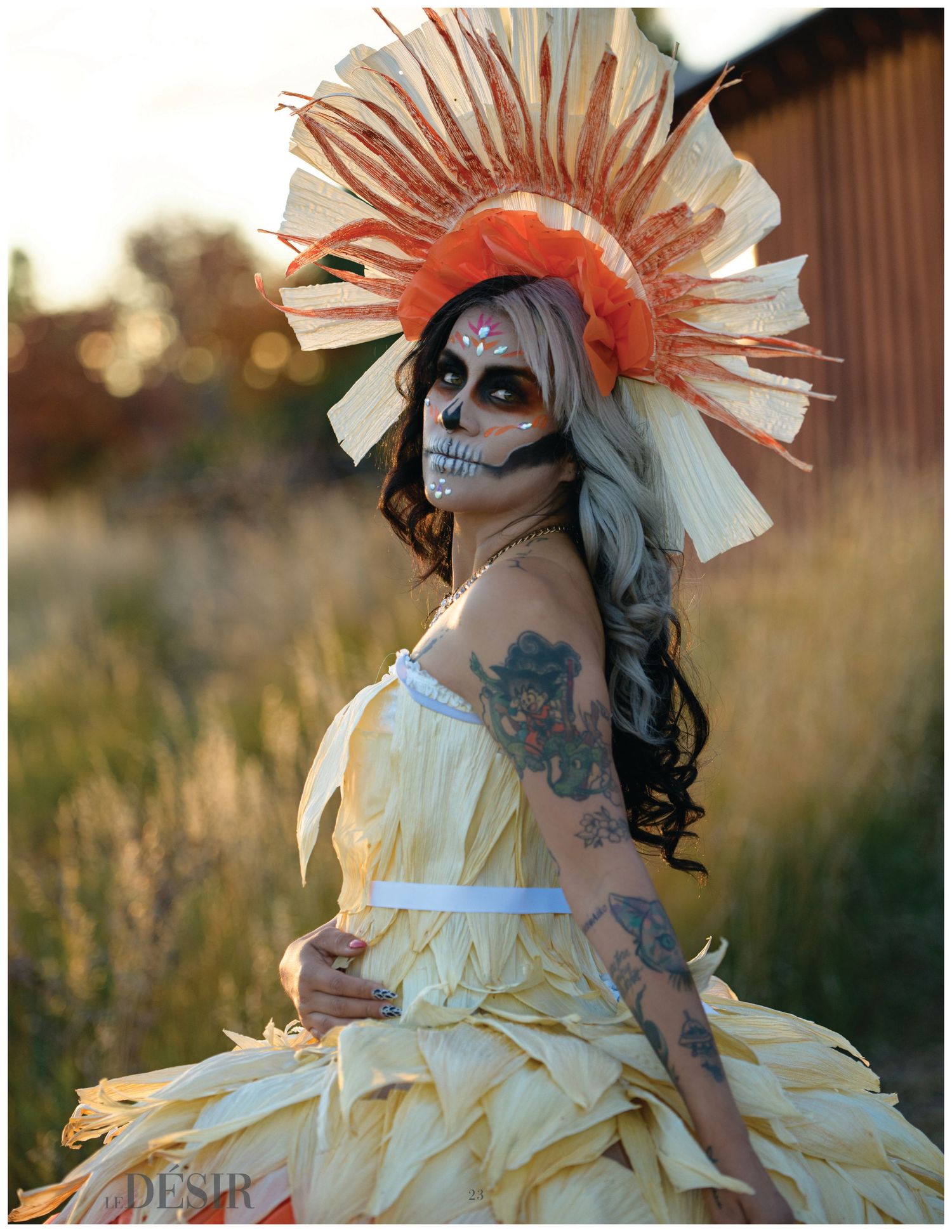 Woman in Day of the Dead skull makeup wears a corn husk dress and a sunburst feathered headdress while posing in a golden field at sunset.