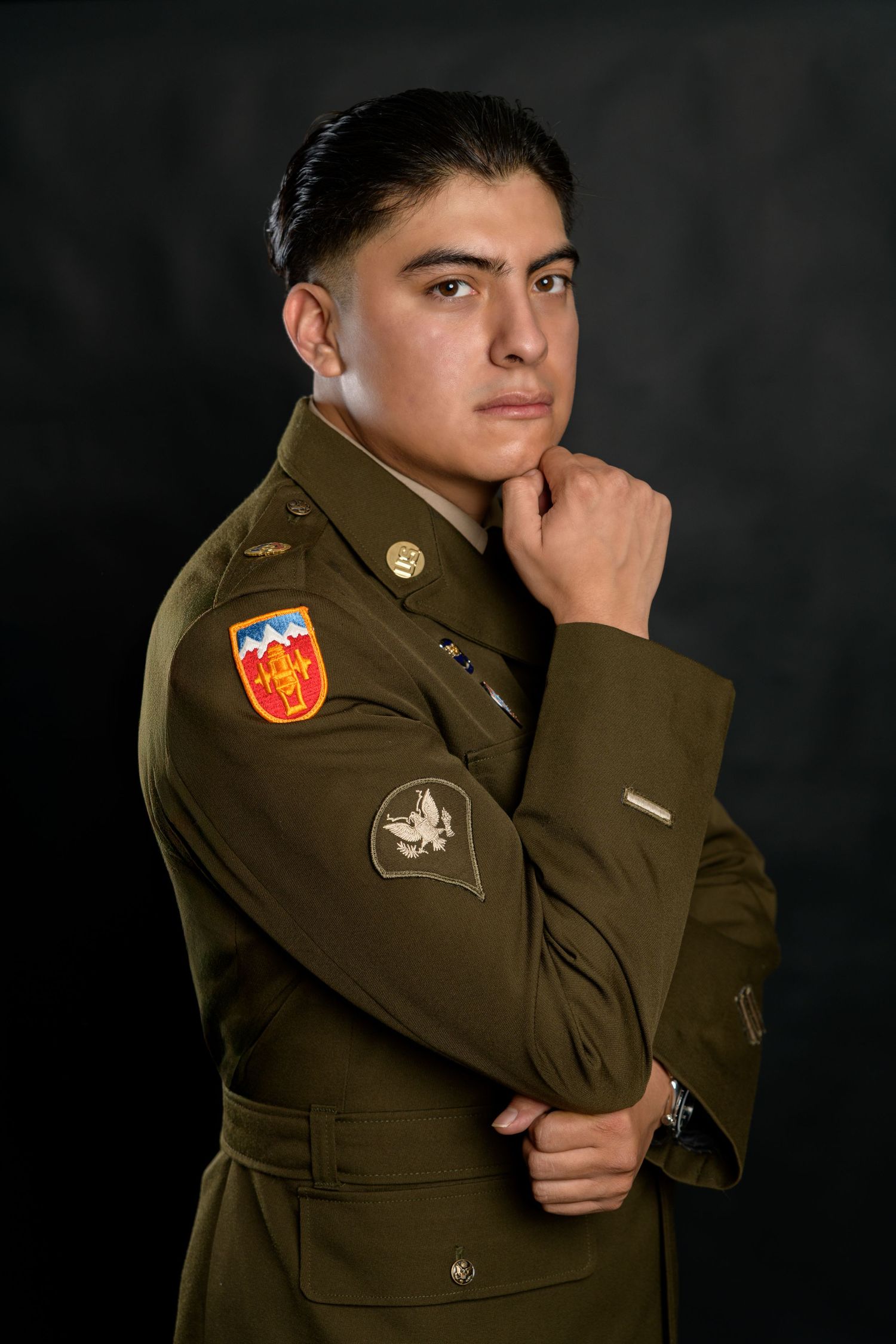 Young man in olive military dress uniform with patches, posing thoughtfully with hand on chin against dark background.