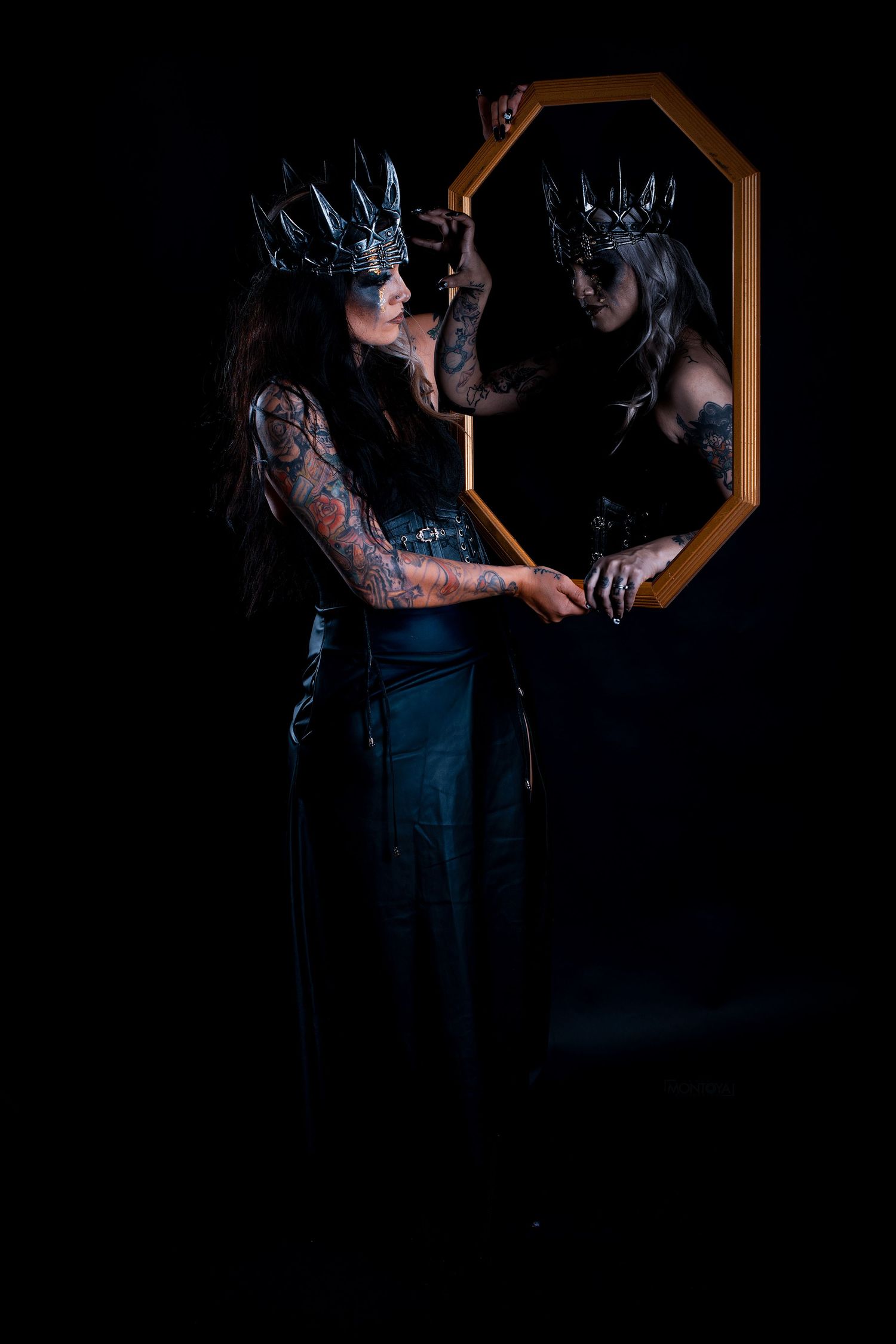Tattooed woman in dark crown and leather outfit gazes into ornate mirror in dramatic dark studio setting.