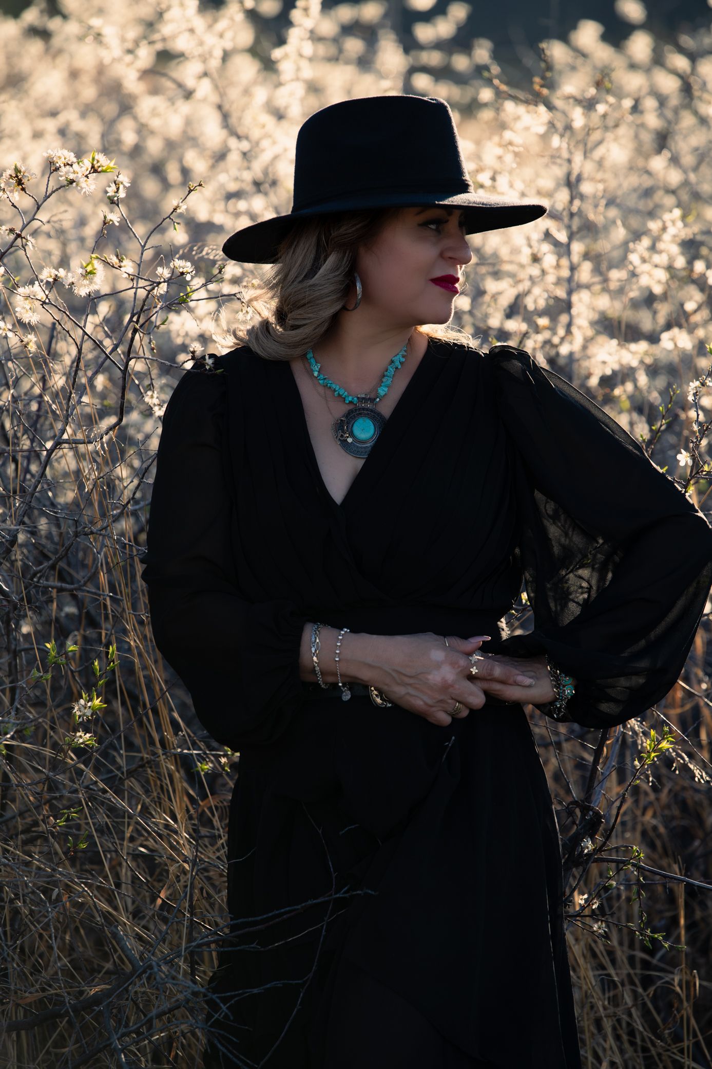 Elegant woman in black wrap dress and wide-brim hat with turquoise necklace standing in sunlit field.