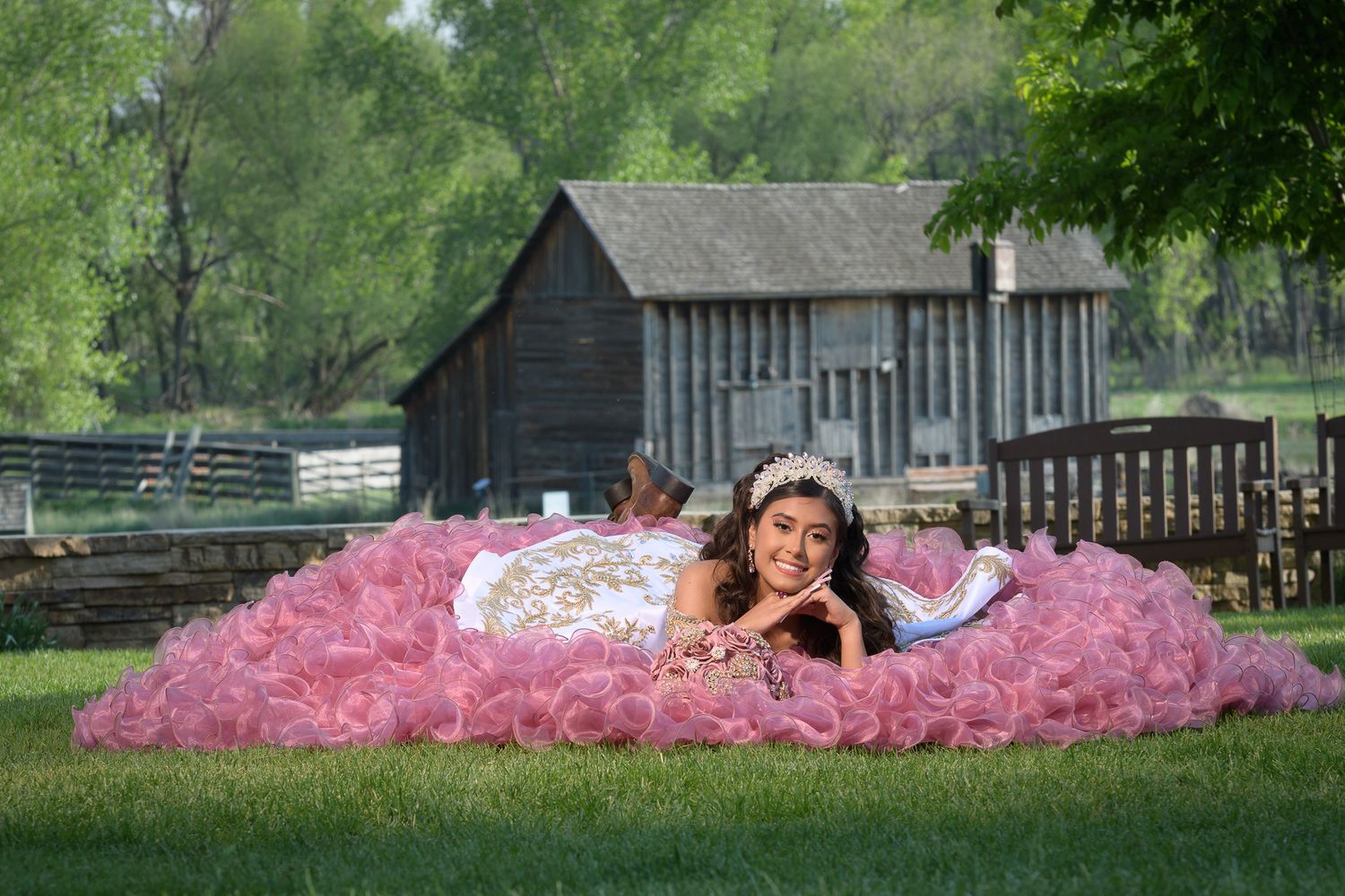 Young woman in pink ruffled quinceañera gown lies on grass with rustic barn in background.