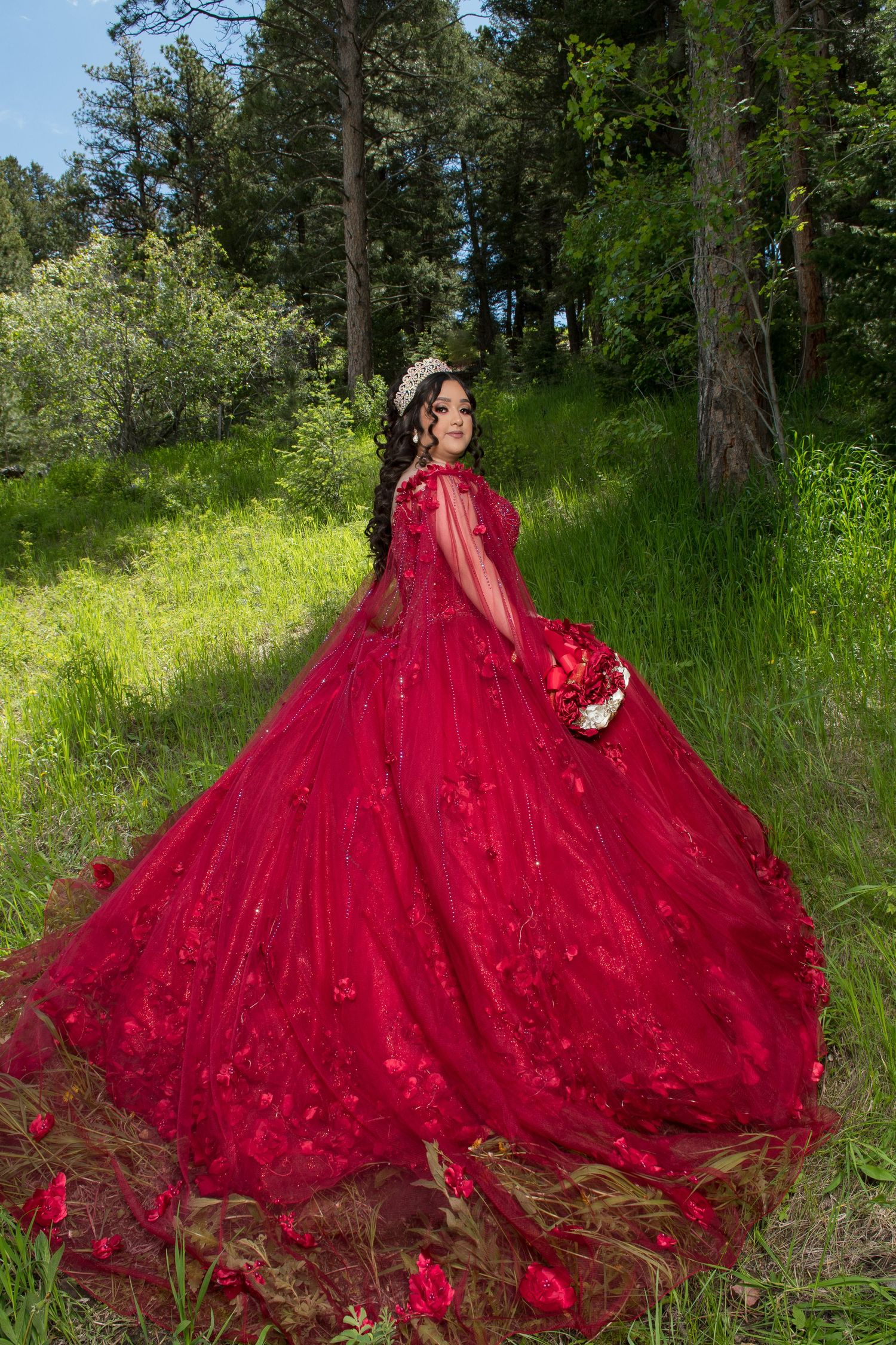 Elegant young woman in red ball gown with floral embroidery and cape twirls in a green forest setting.
