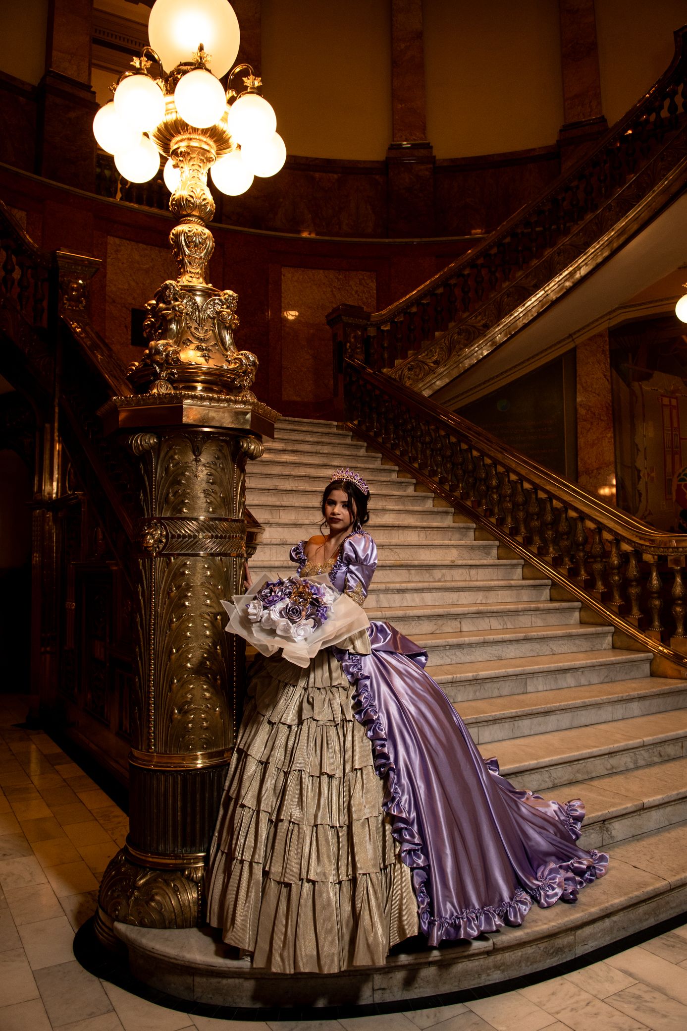 Woman in elegant purple and gold tiered quinceañera gown standing by ornate lamp on marble staircase.