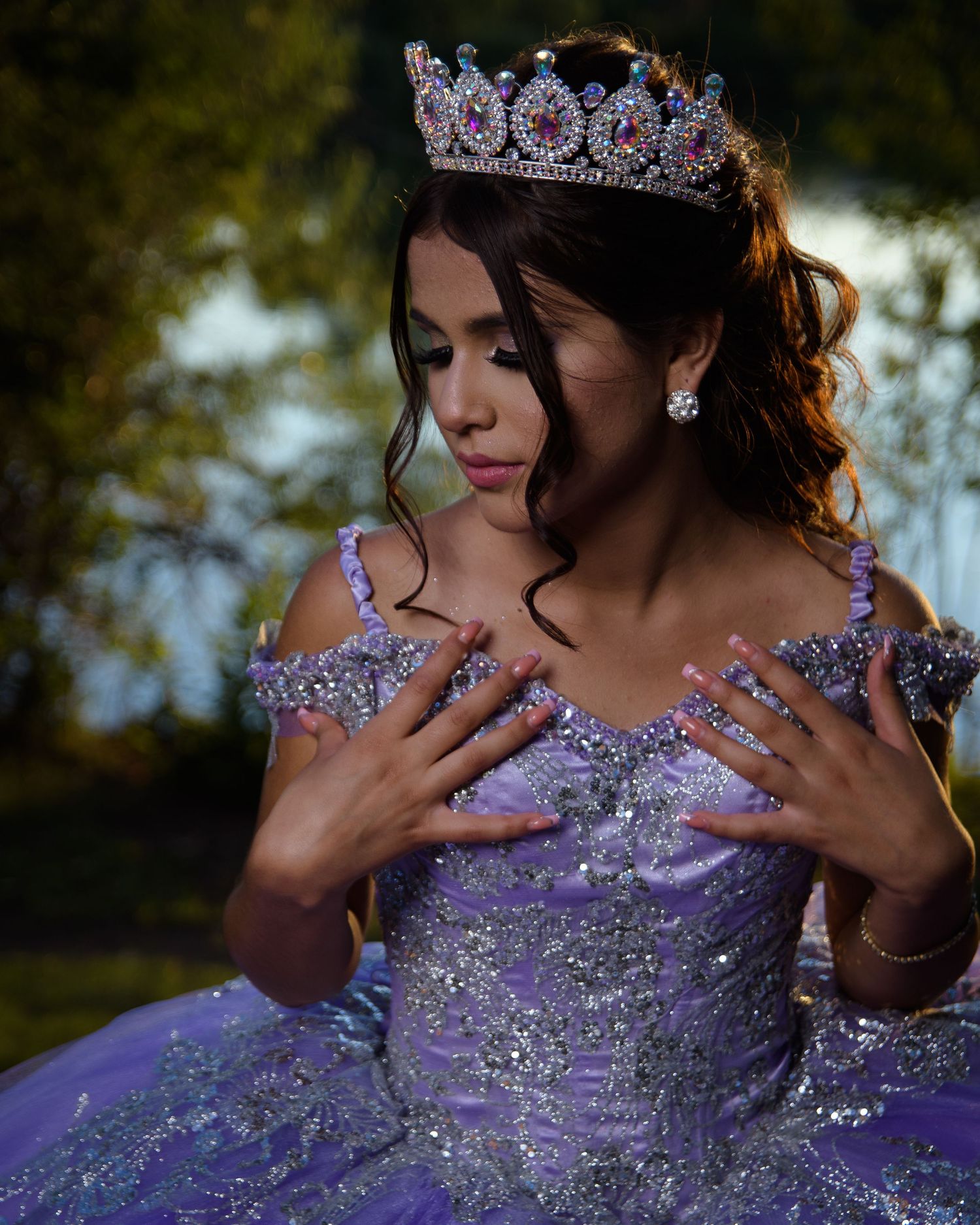 Young woman in a sparkling lavender quinceañera ball gown and crown poses outdoors at sunset.