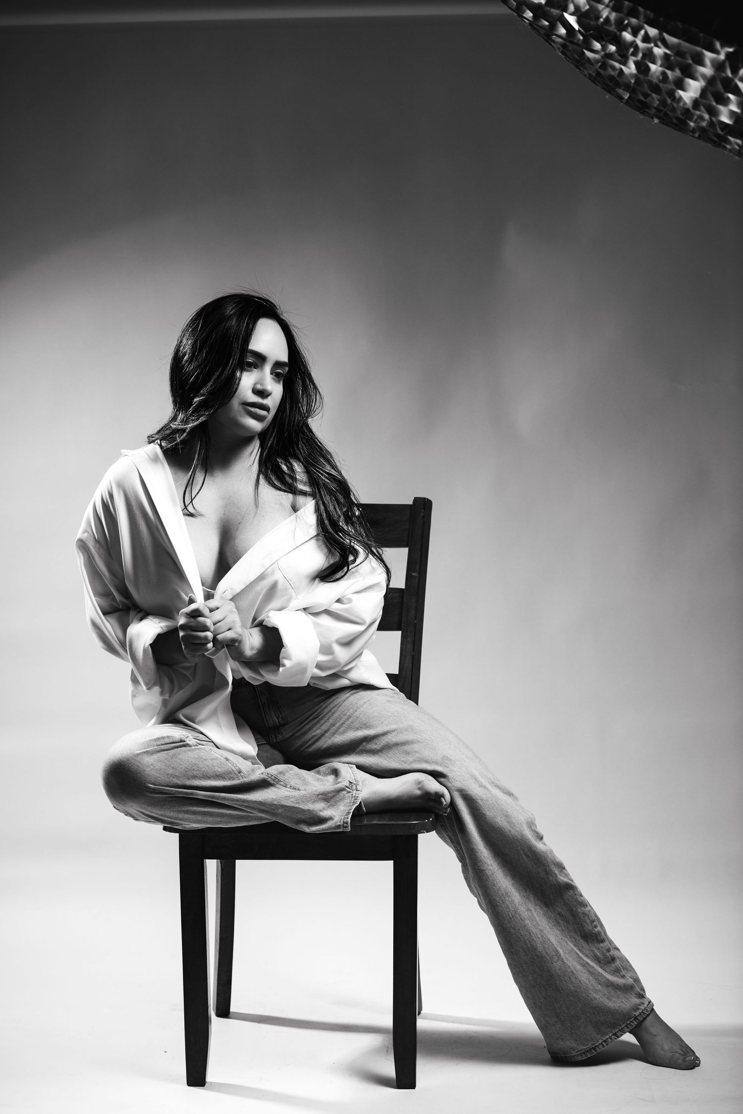 Black and white studio photo of a woman in an open shirt and wide-leg pants seated on a wooden chair.