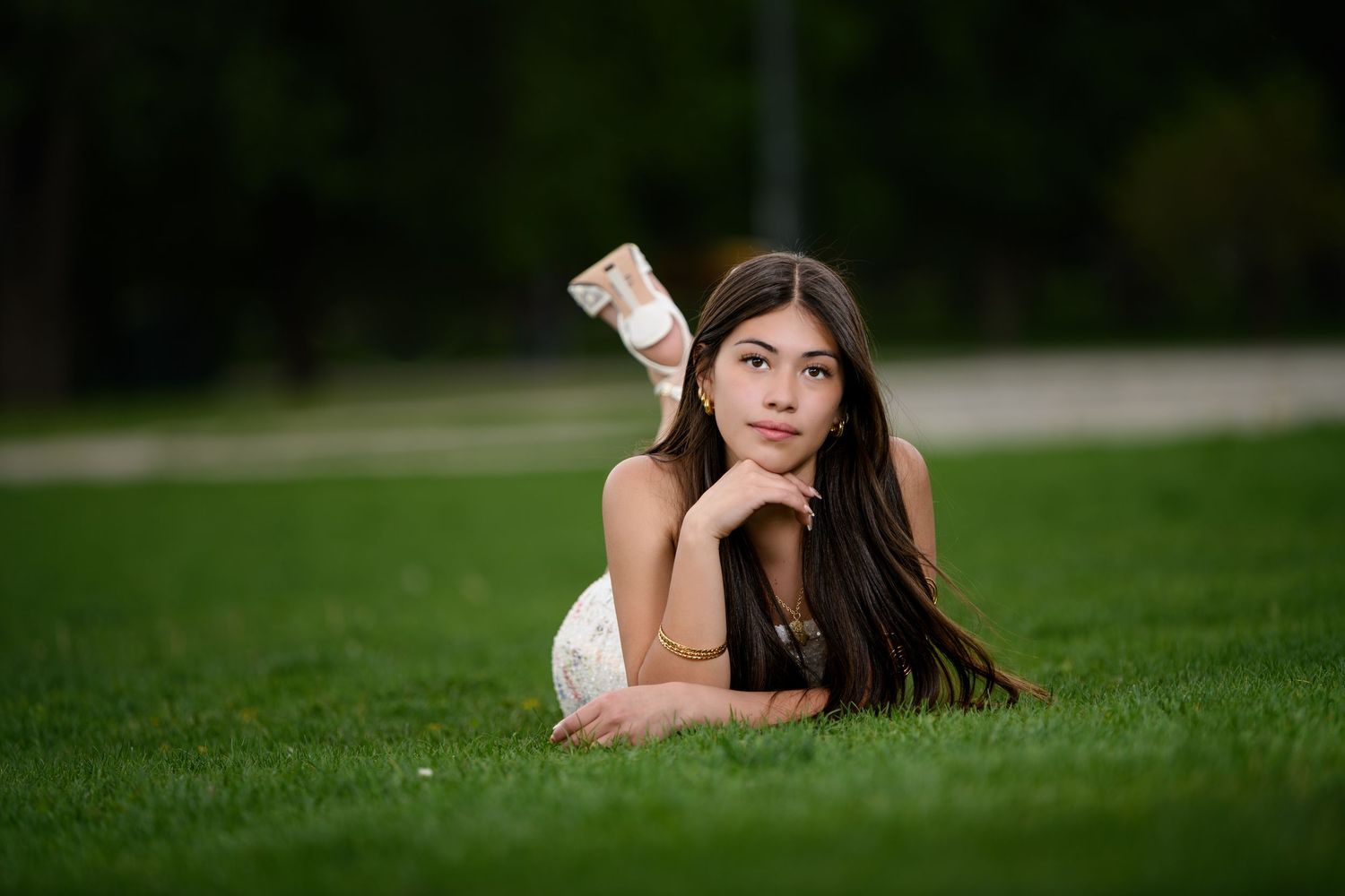 Young woman in white dress lying on green grass in a park, resting chin on hands.
