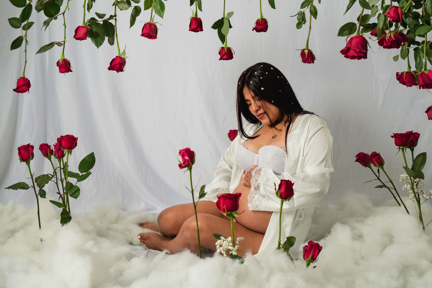 Pregnant woman in white robe sits among fluffy clouds with red roses hanging on white backdrop.