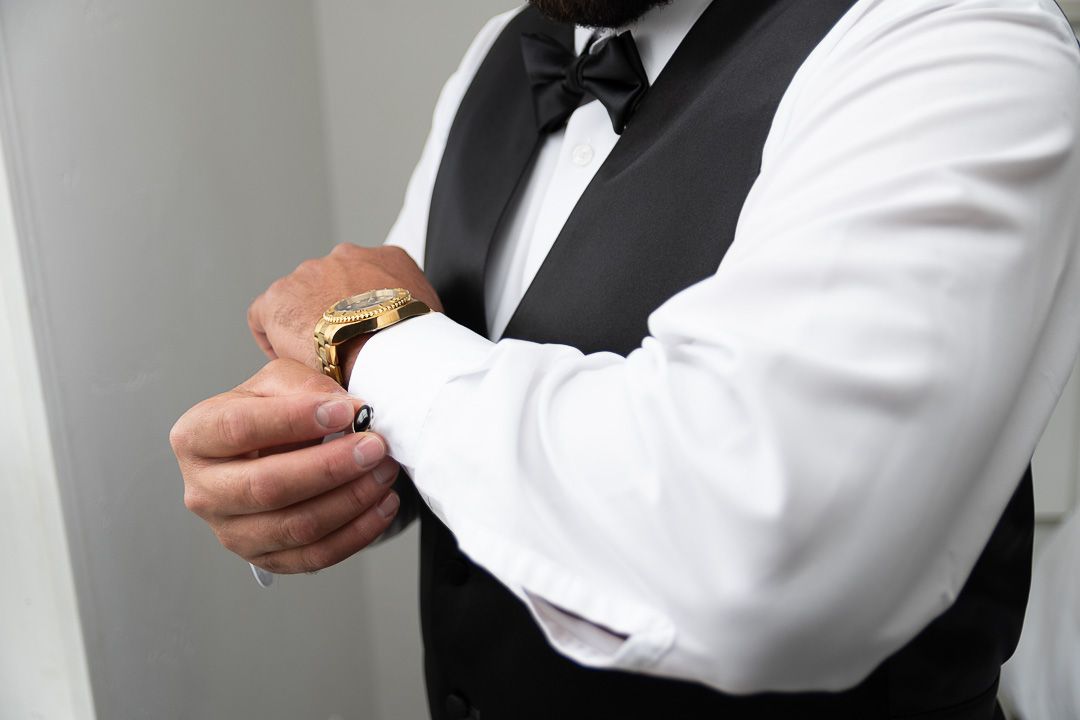 Groom adjusting gold cufflinks on white dress shirt with black bow tie and vest.