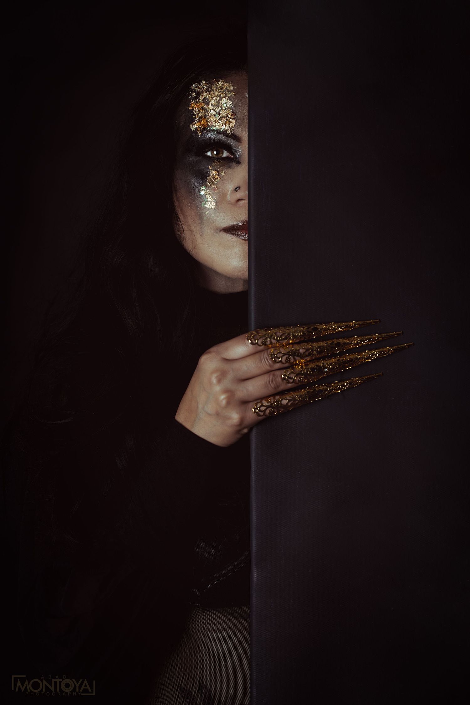 Mysterious woman with gold glitter makeup and long gold nails peers dramatically around dark wall.