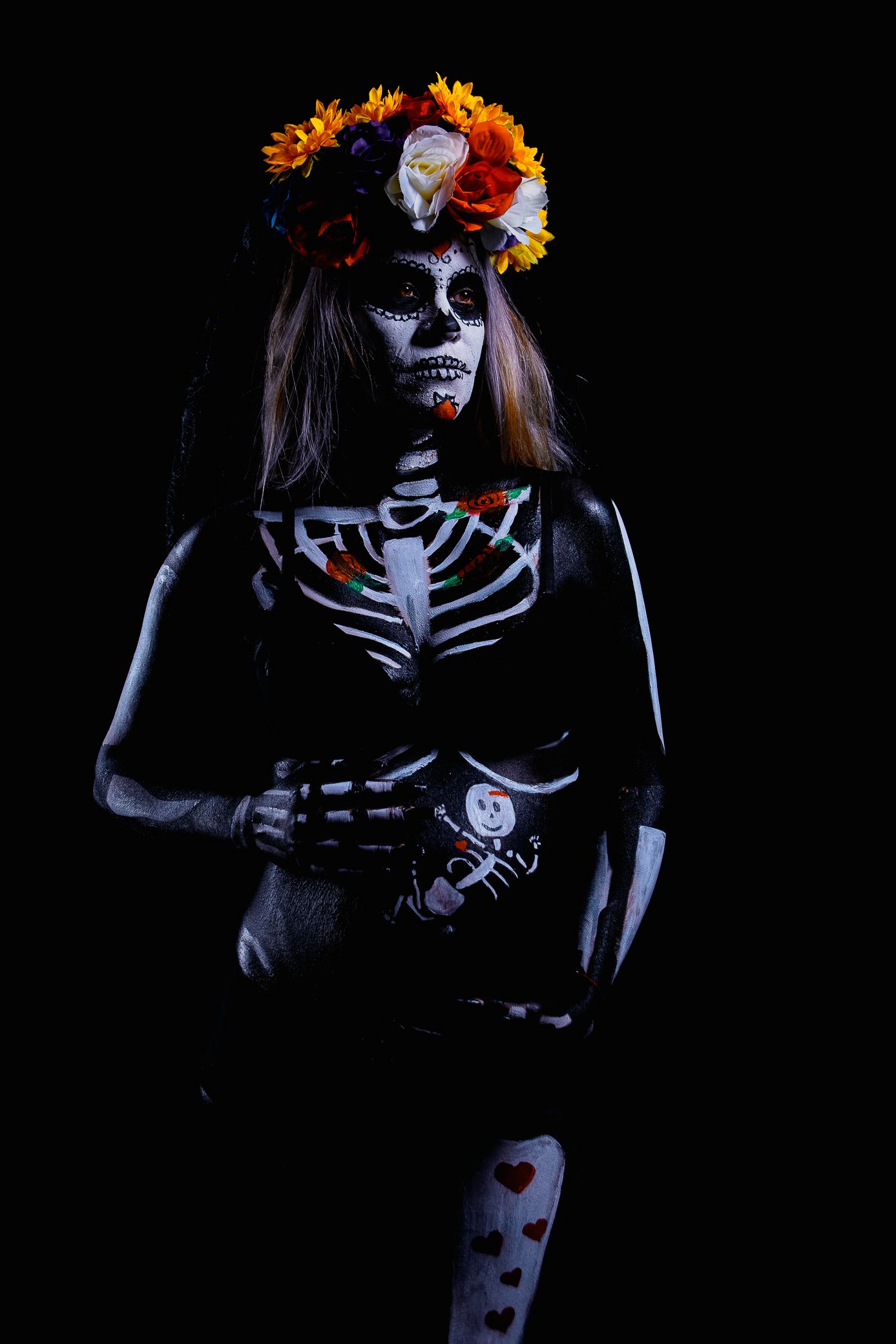 Woman in dramatic Day of the Dead skeleton body paint and floral crown poses on dark background.
