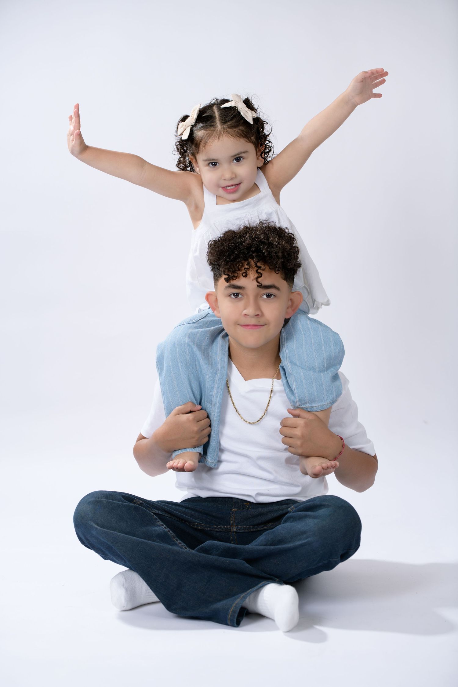 A boy sits cross-legged with a young girl on his shoulders, both smiling, wearing casual white and denim outfits.