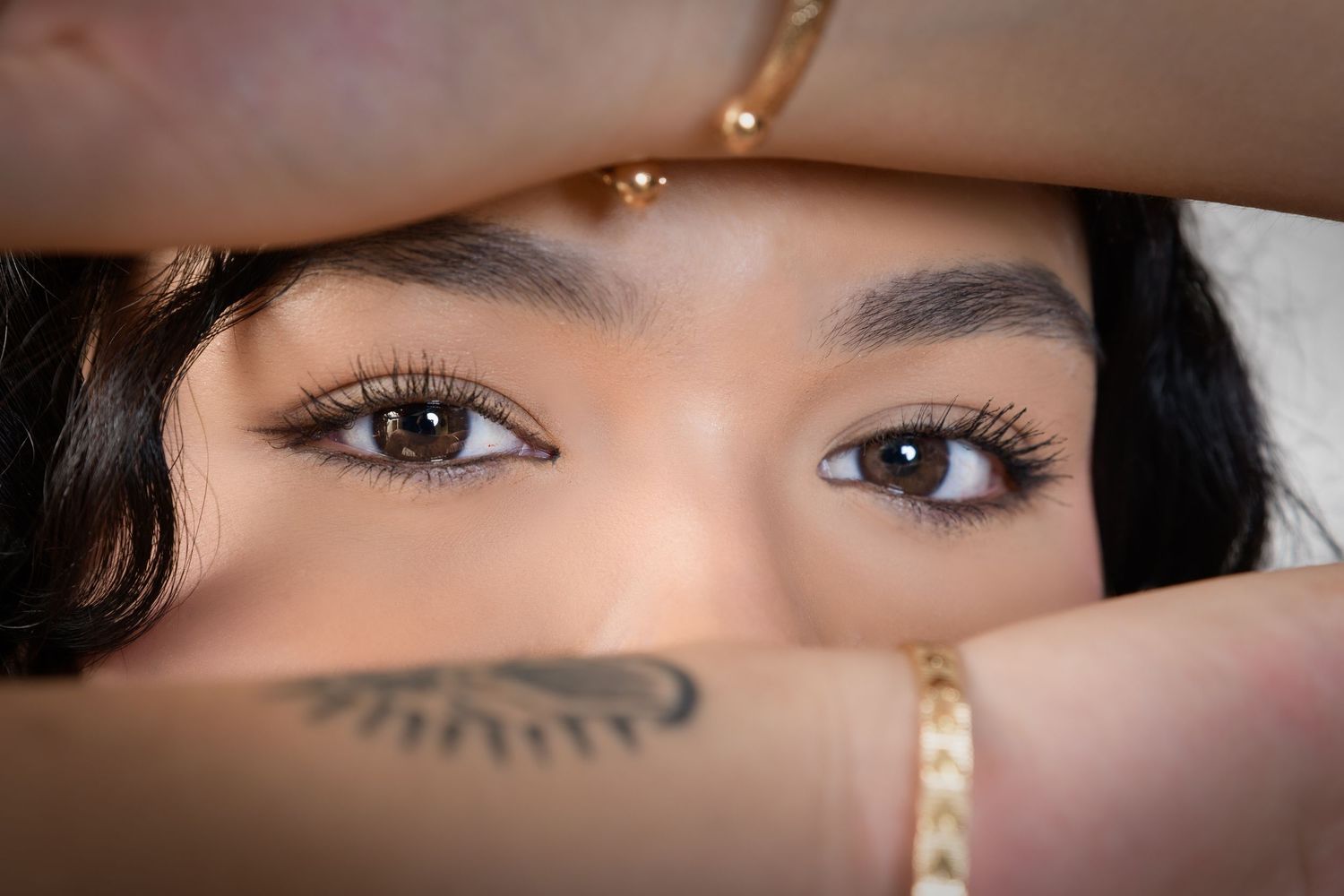 Close-up portrait of young woman with brown eyes framed by hands adorned with gold jewelry and tattoos.