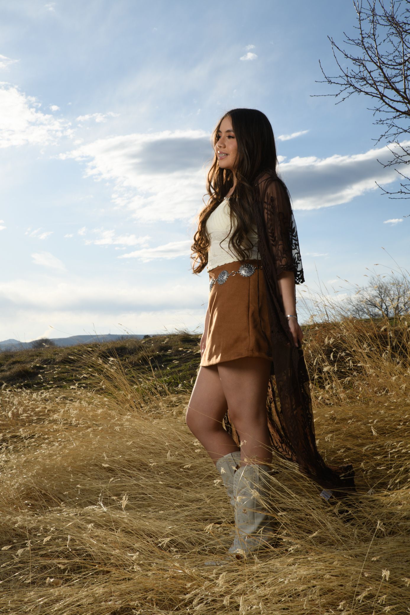 Woman in boho western outfit with fringe, mini skirt, and cowboy boots standing in golden field.