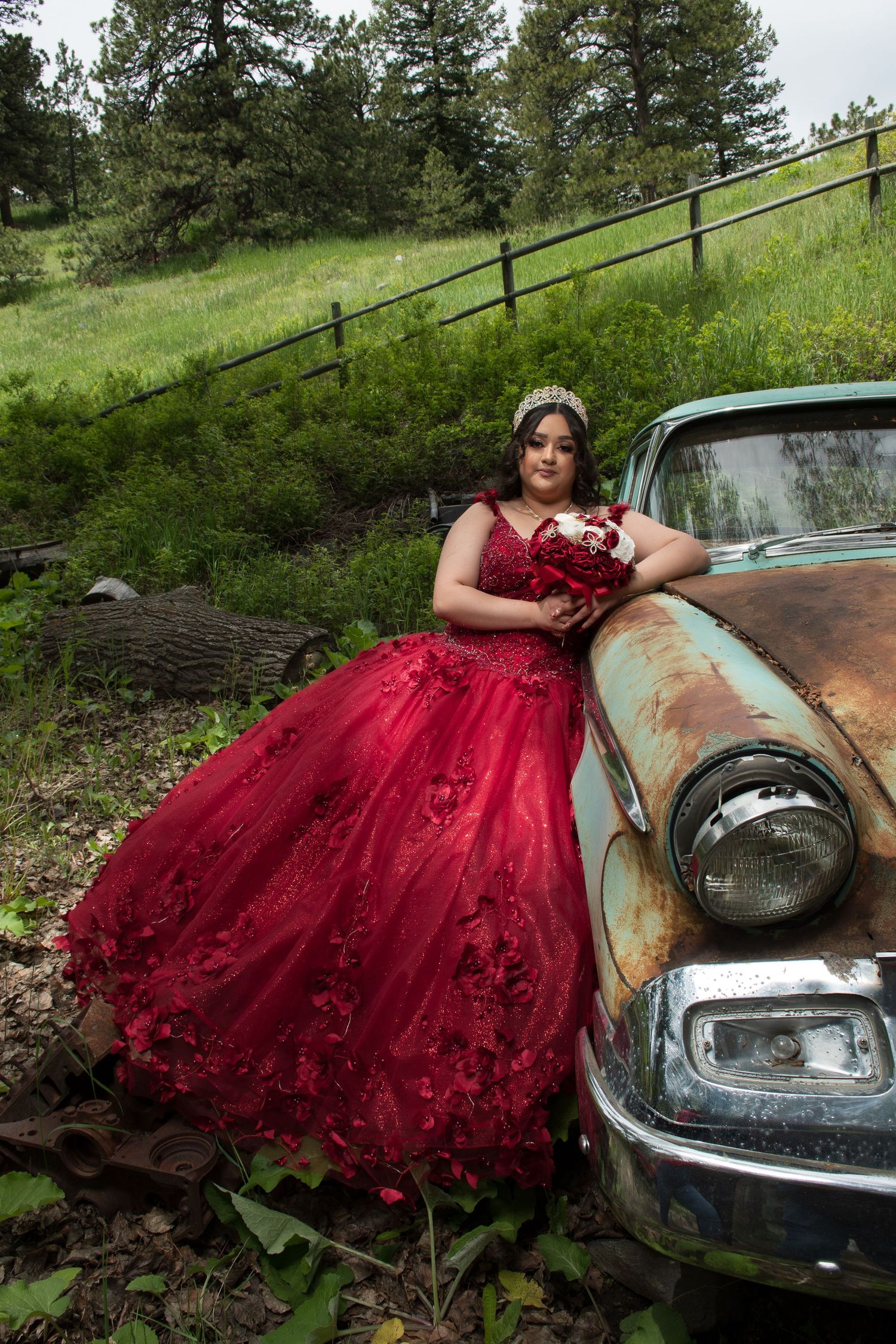 Young woman in red quinceañera dress with tiara leans against rustic vintage car in outdoor setting.