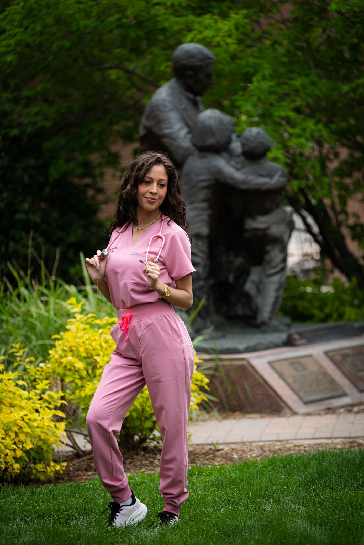 Woman in pink medical scrubs with stethoscope posing near bronze statue in garden.