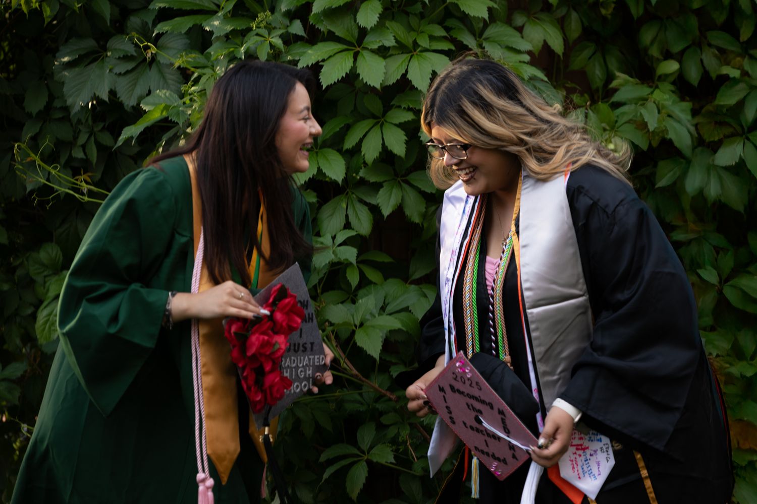 Two female graduates in caps and gowns laughing together in front of green ivy wall outdoors.