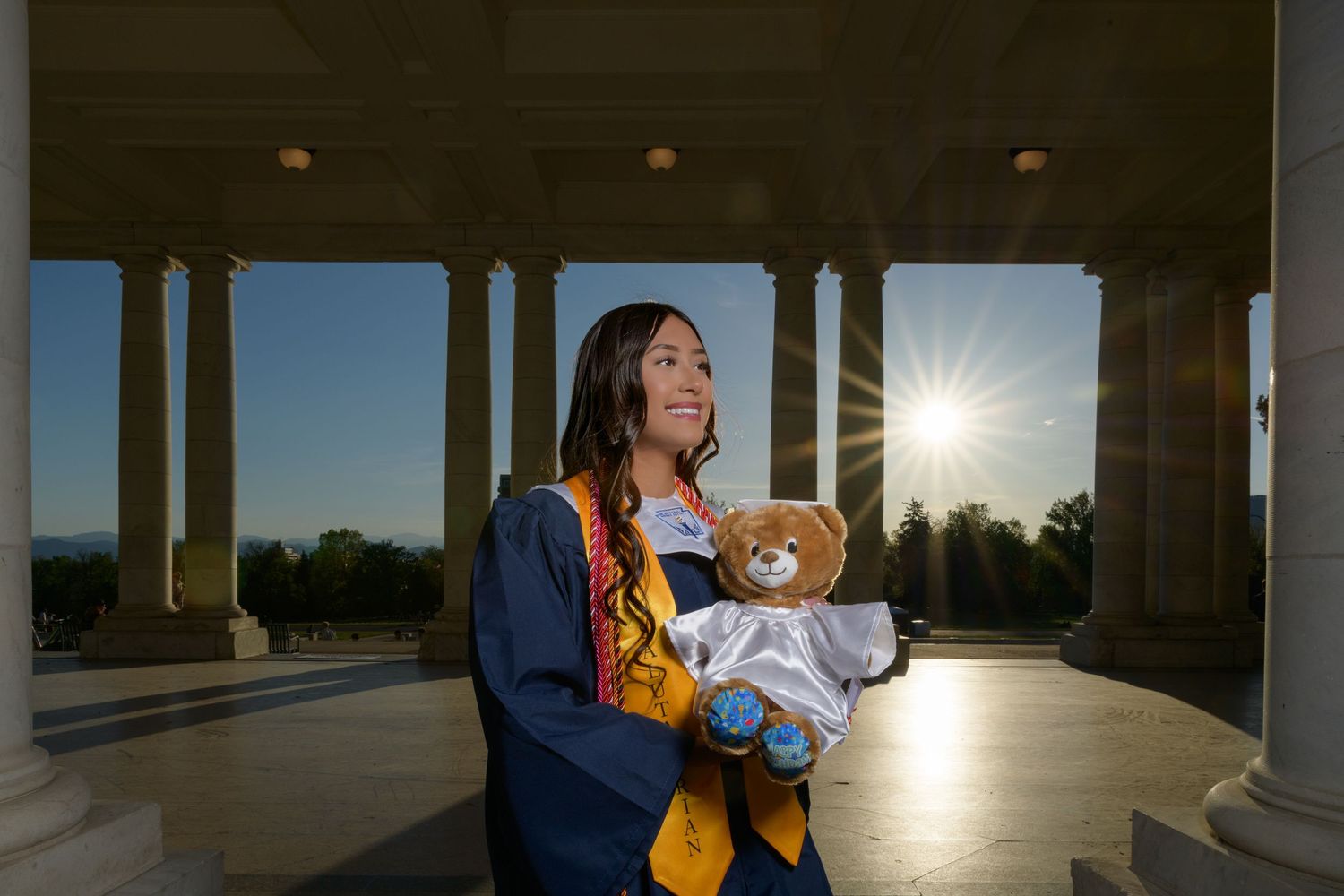 Female graduate in blue and gold gown holding teddy bear at sunset colonnade with sunburst.