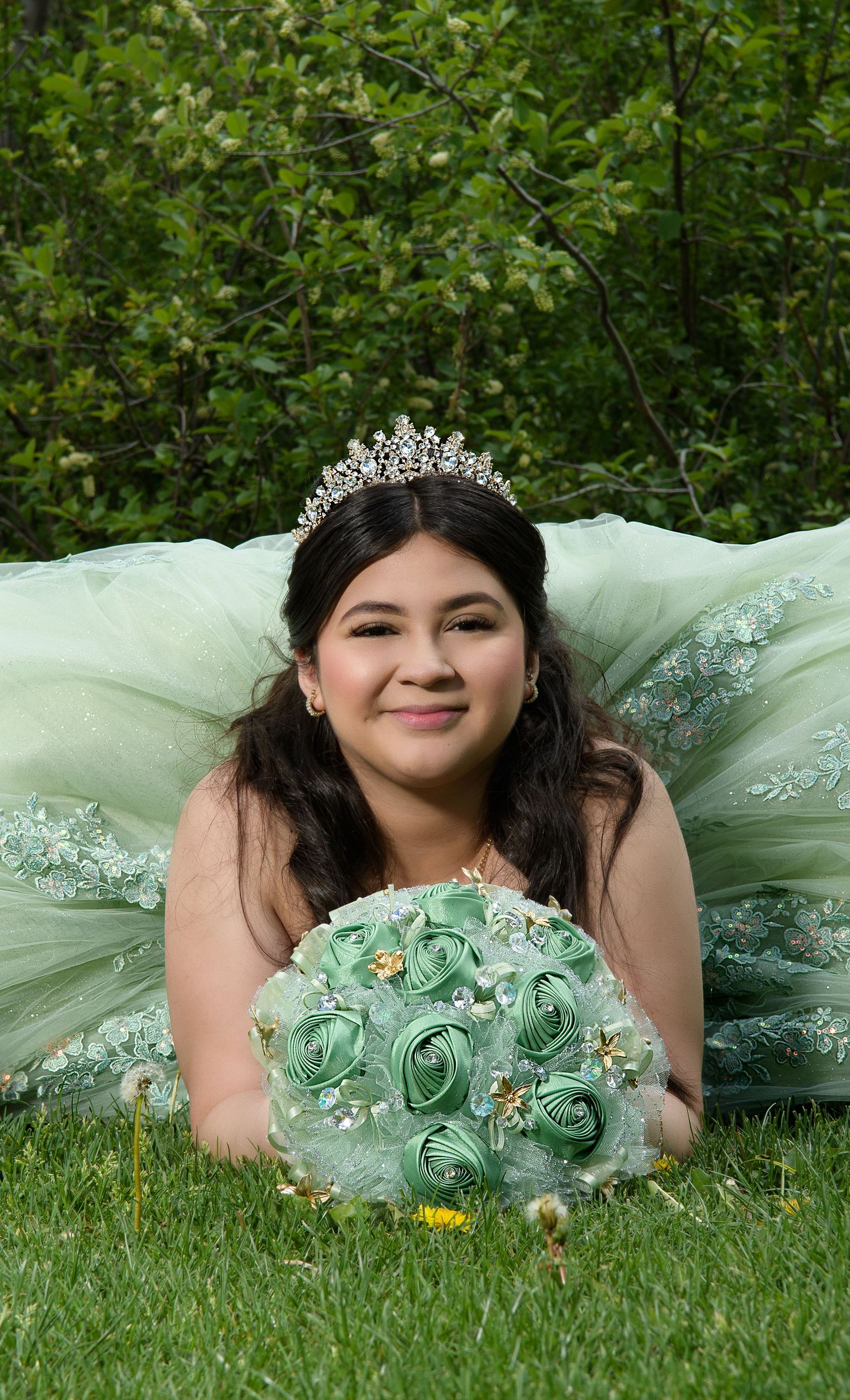 Smiling young woman in mint green quinceañera dress with tiara lying on grass holding rose bouquet.