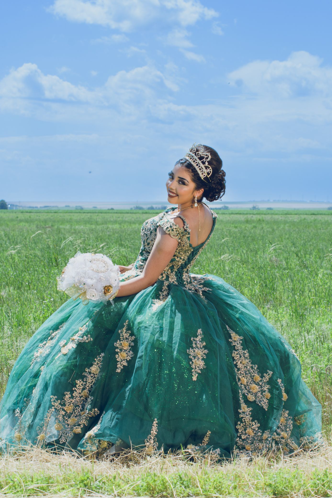 Smiling young woman in teal green quinceañera gown with gold embroidery poses in open green field.