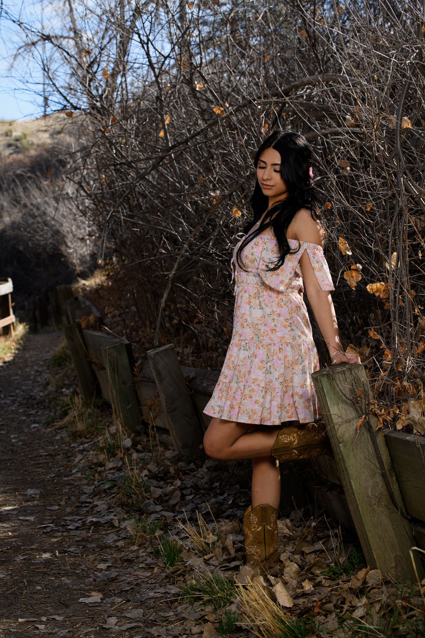 Young woman in floral dress and cowboy boots leaning on wooden fence along autumn nature trail.