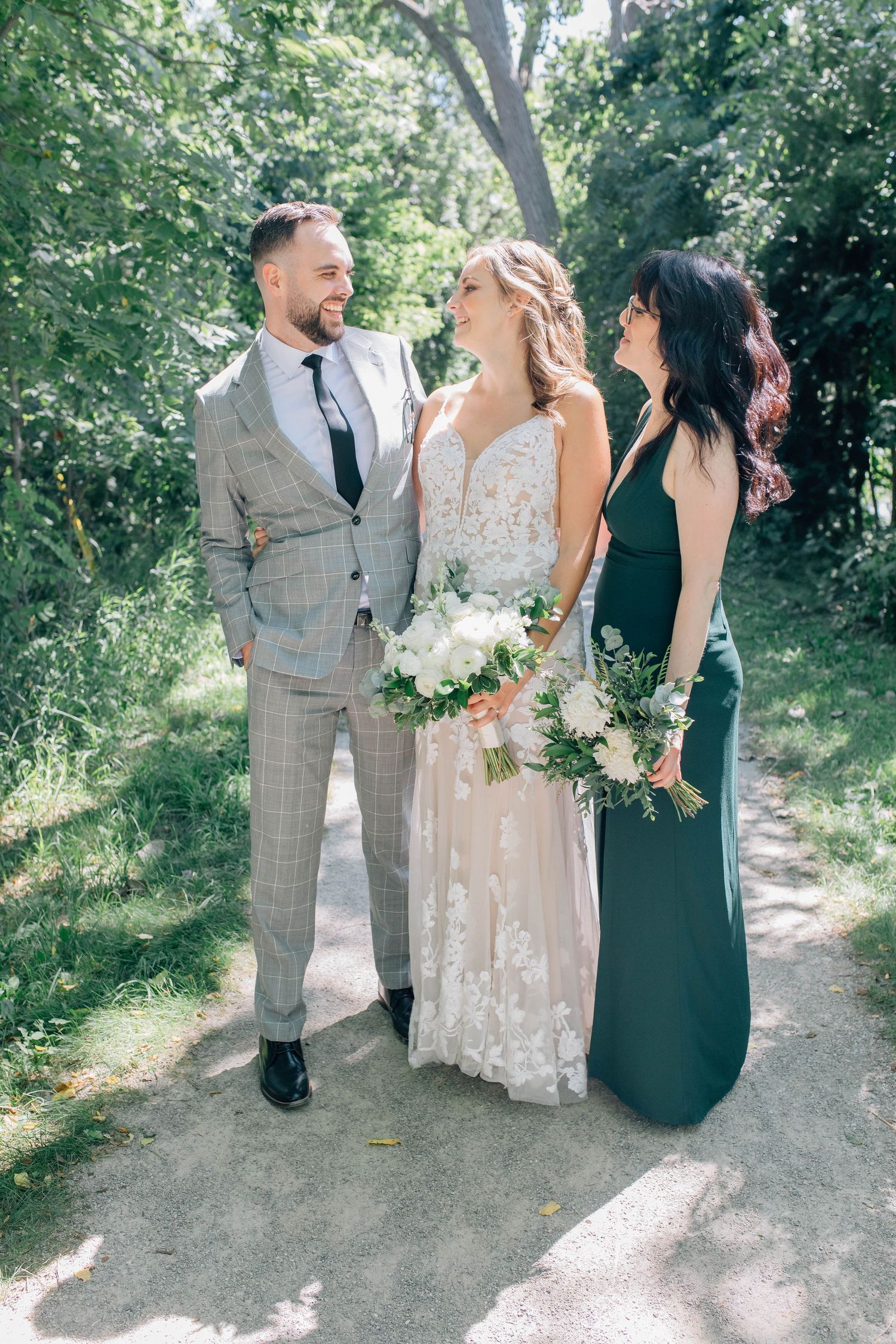 Bride in lace gown stands between groom in plaid suit and bridesmaid in emerald dress on a garden path.